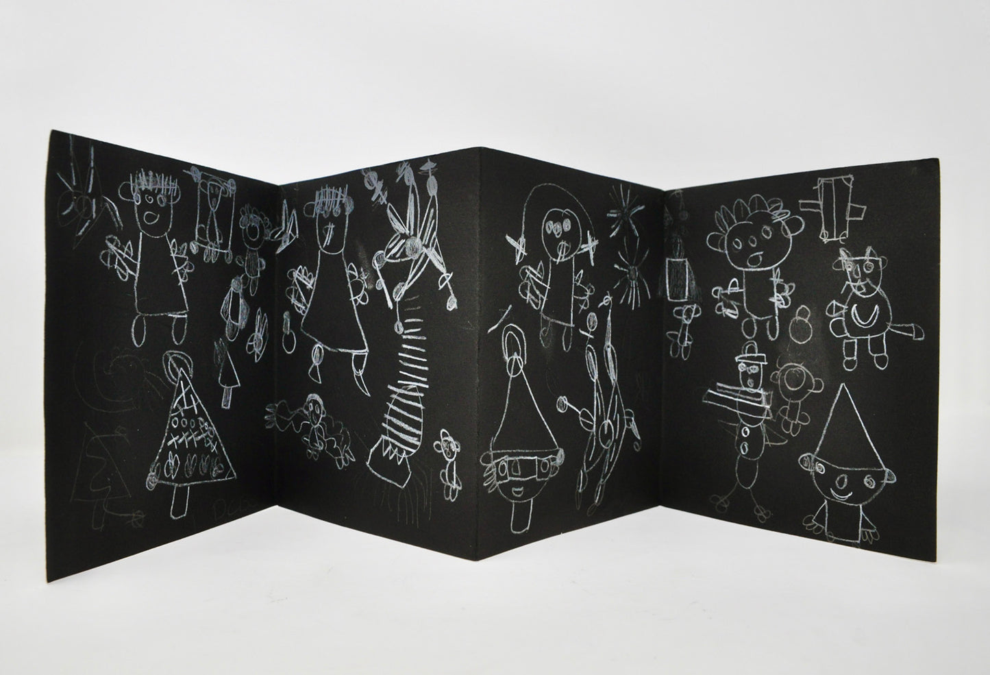 One side of an accordion book made with black paper, photographed on a white backdrop. Using white pencil, the artist has filled the surface with drawings of stylized figures and striped symbols. The figures have large round heads, dashes or petals representing hair, tall triangle hats, and rectangular bodies. Other drawn forms include trees, cats, star bursts, and flowers.
