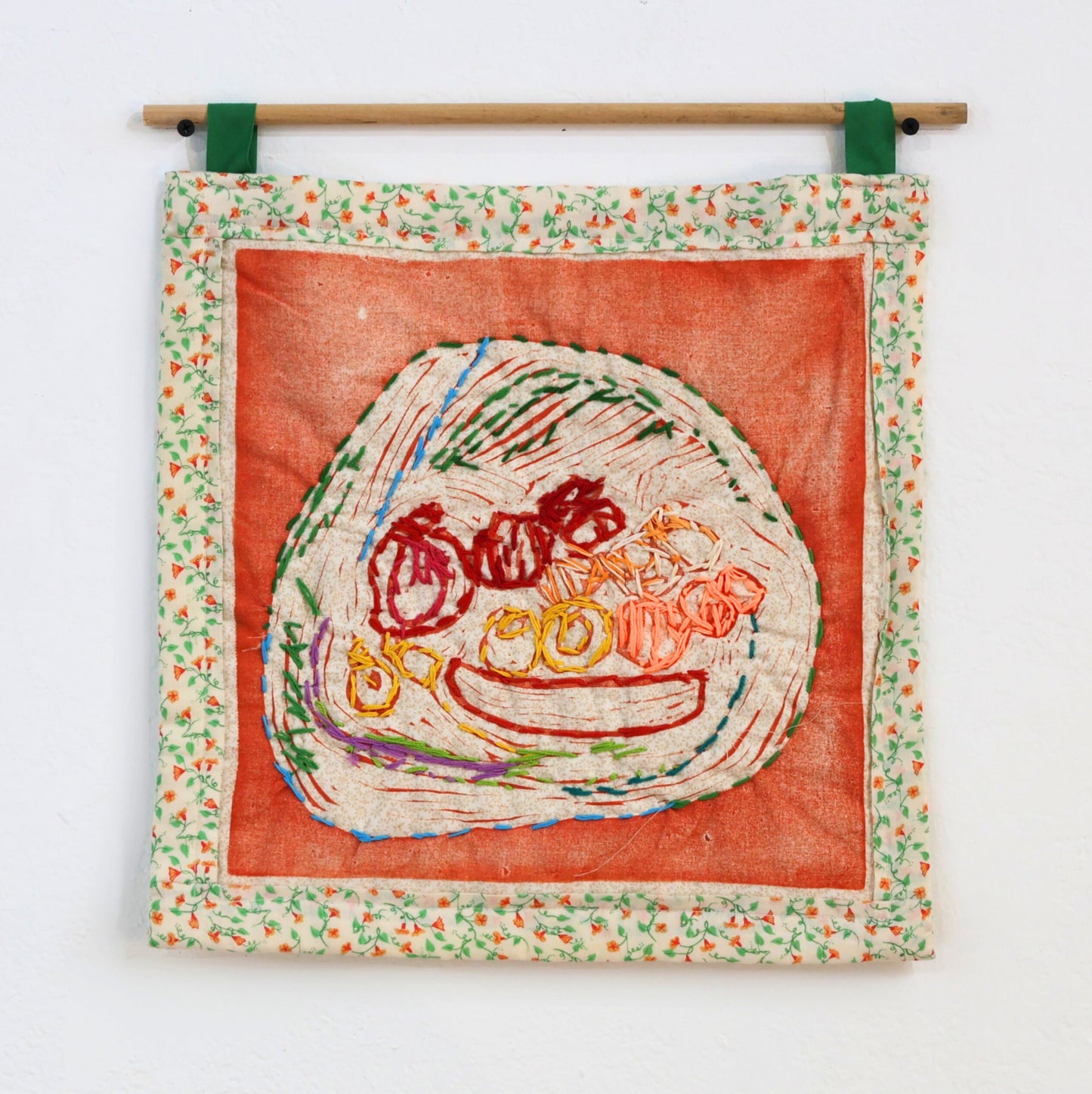 A small square wall hanging textile with colorful embroidery representing a bowl of fruit. The embroidery is made inside of a white circle on an otherwise orange square of fabric. There is a floral patterned border, and the piece hangs by two green loops from a wooden dowel.