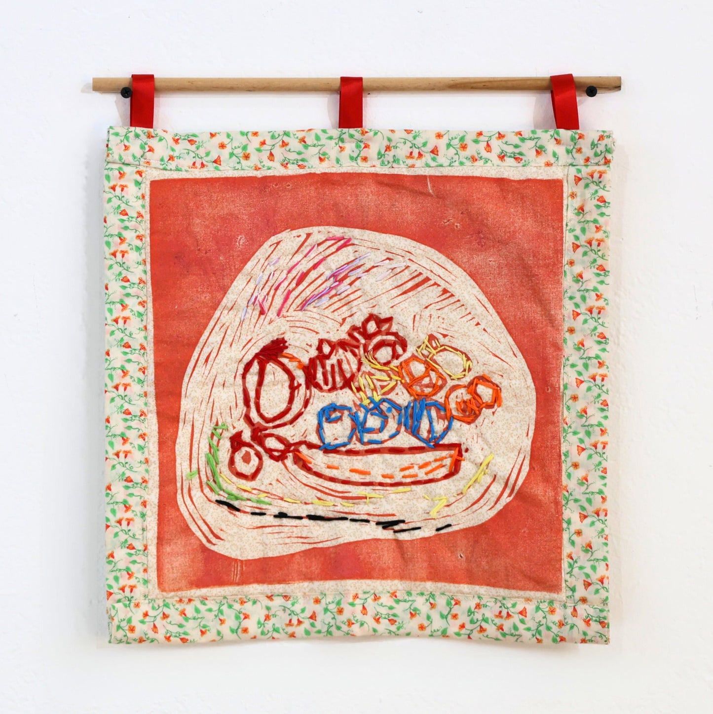 A square wall hanging of an embroidered bowl of fruit, with a floral border, hung with red tabs on a wooden rod.
