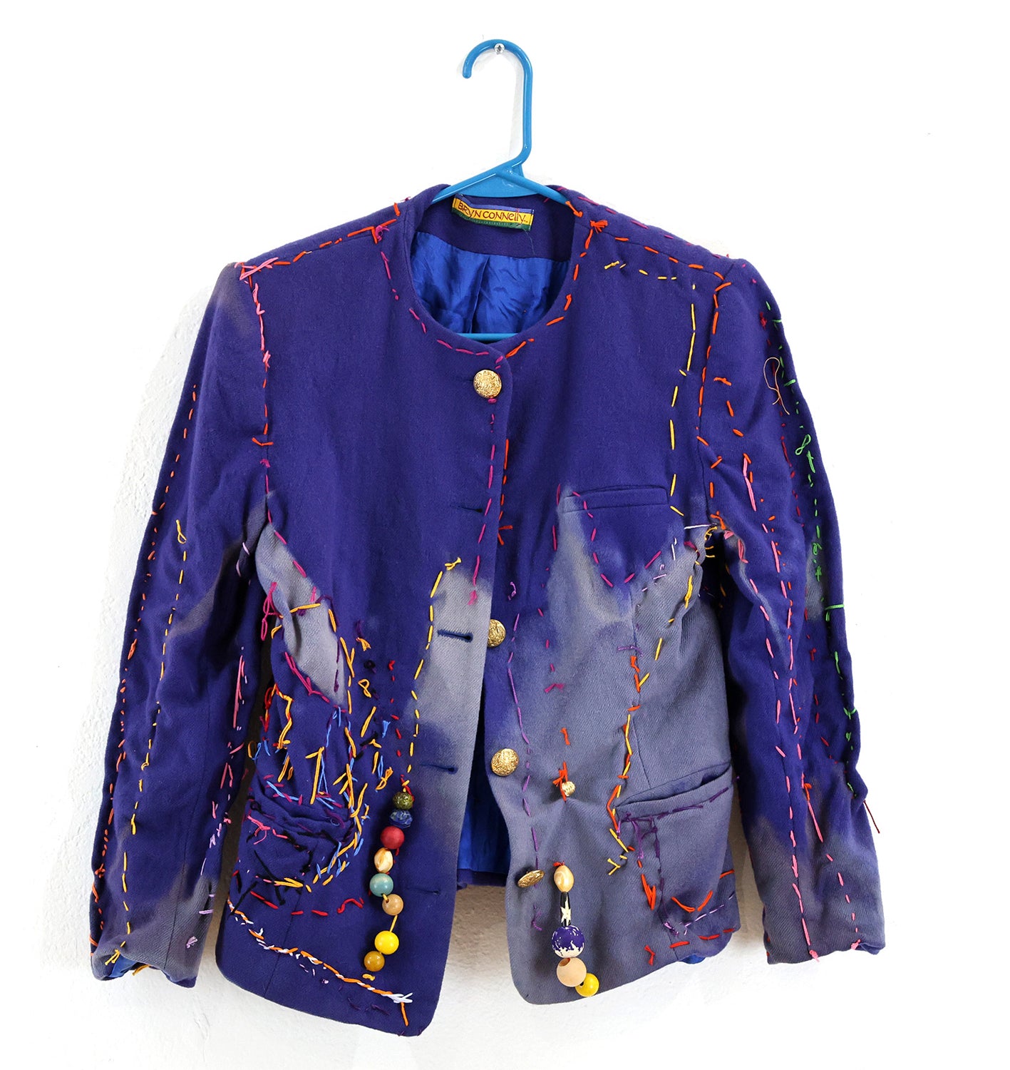 Purple wool jacket bleached by the sun in a light purple color across the center-bottom-right of the jacket. The jacket has golden buttons and is adorned with colorful beads and stitching in red, pink, yellow, blue and green colors. The stitching mainly covers the silhouette of the jacket and the bottom-center-left of the jacket in a swirl of colors. The jacket hangs on a blue clothes hanger against a white background.