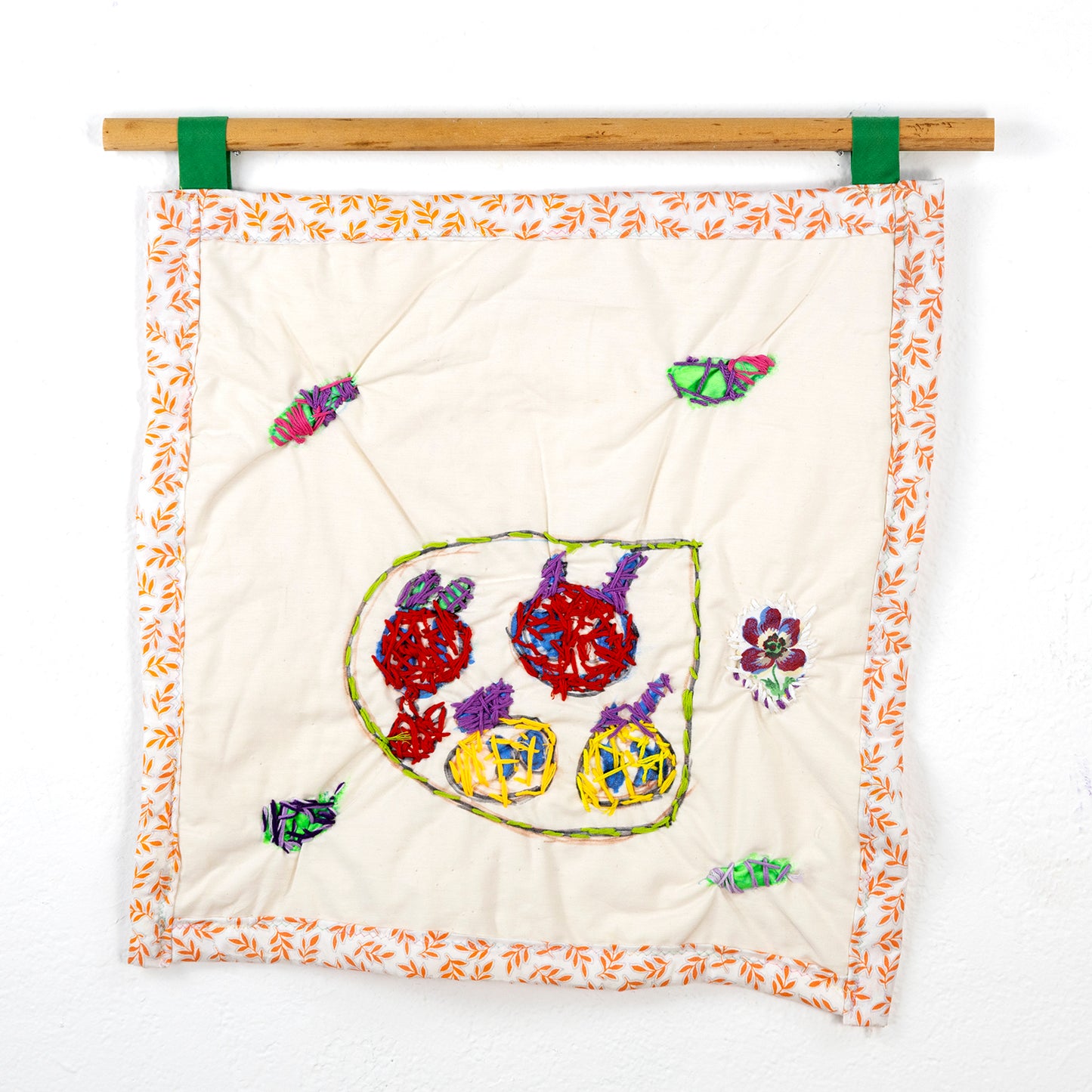 A small square quilt that hangs from a wooden rod with green loops. The fabric is off-white with an orange and yellow leaf-patterned border. Centered on the fabric is an abstract embroidery piece featuring various colored shapes within a green-bordered area. There are red, purple, and yellow circular shapes with textured stitching. Scattered around this central design are small embroidered elements, including colorful leaf-like and floral shapes.