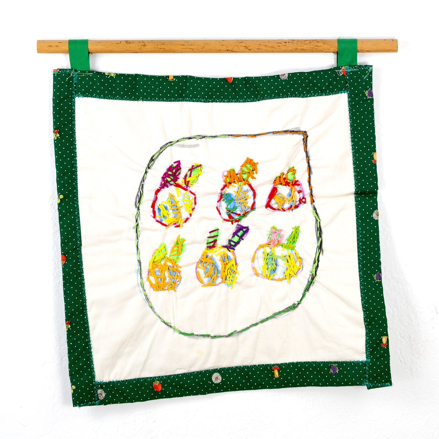 A small square quilt that hangs from a wooden rod with green loops. The quilt features a central design of six stylized fruit-like objects, each crafted with multicolored threads and outlined with vibrant stitching. The edge of the piece has a green border with small white polka dots, adorned with occasional fruit and flower motifs.