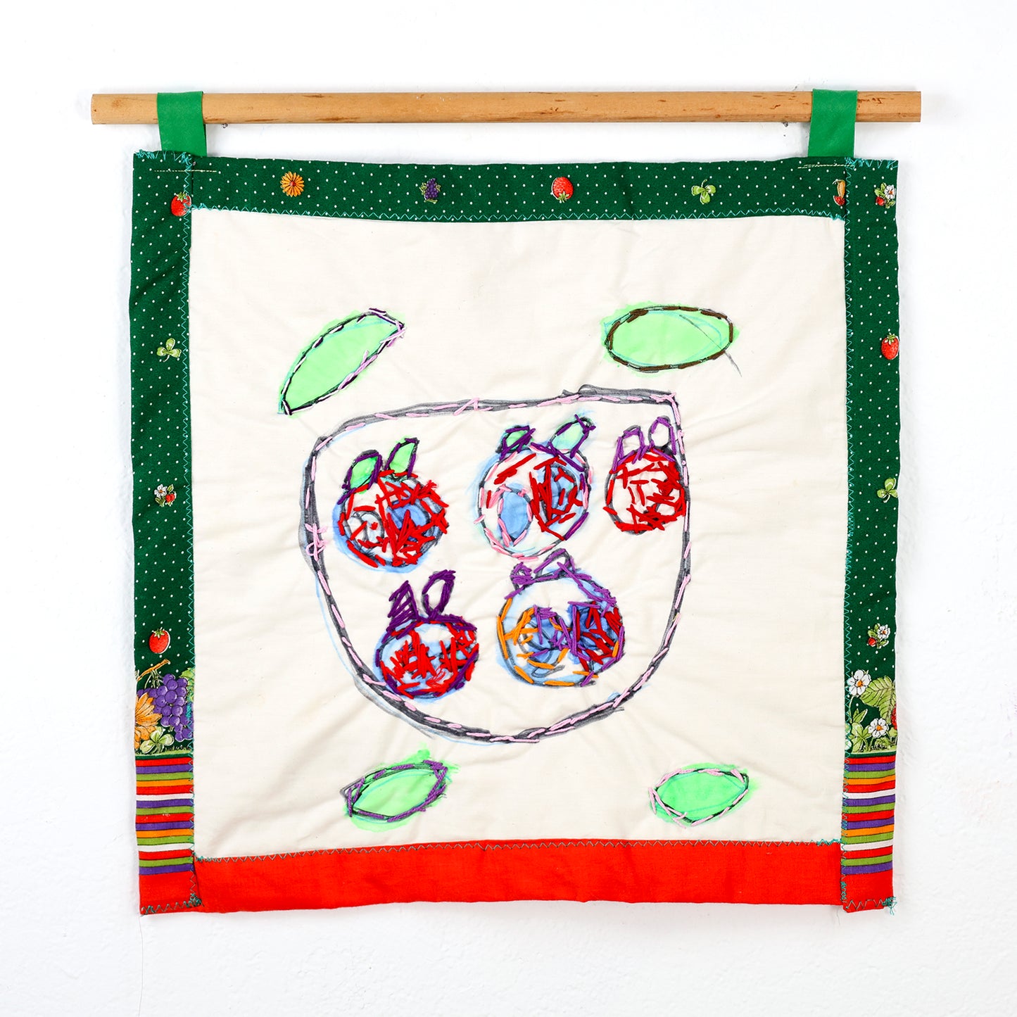 A small square quilt that hangs from a wooden rod with green loops. The centerpiece is a cream white fabric with embroidery of five abstract fruit shapes, contained within a stylized bowl formed by a purple outline. Surrounding the central bowl are four oval shapes with light green embroidery, resembling leaves. The bottom edge of the fabric is bordered with bright red, while the lower left corner displays a narrow strip of multicolored stripes.