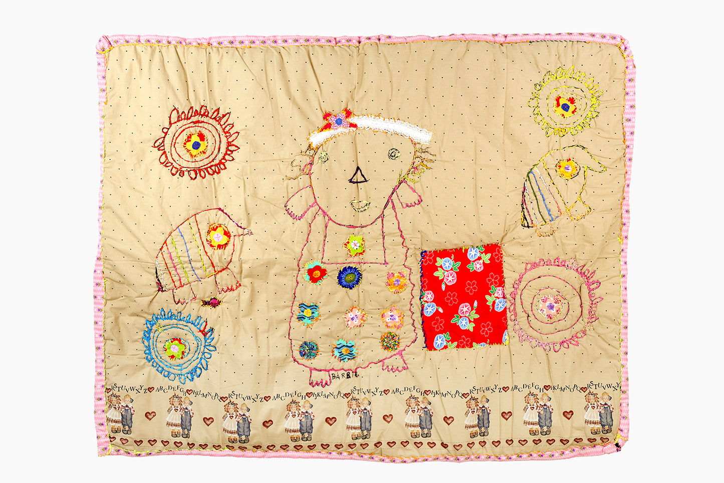 A quilt with a beige background adorned with multicolored embroidery. Central to the design is a figure, outlined in pink thread, decorated with colorful floral patches on the clothes. Surrounding the figure are round flowers and birds, crafted with red and blue embroidery. A notable square patch with red floral fabric is sewn onto the quilt. The border is pink, and the bottom edge features the word "Barbie" written in black marker.