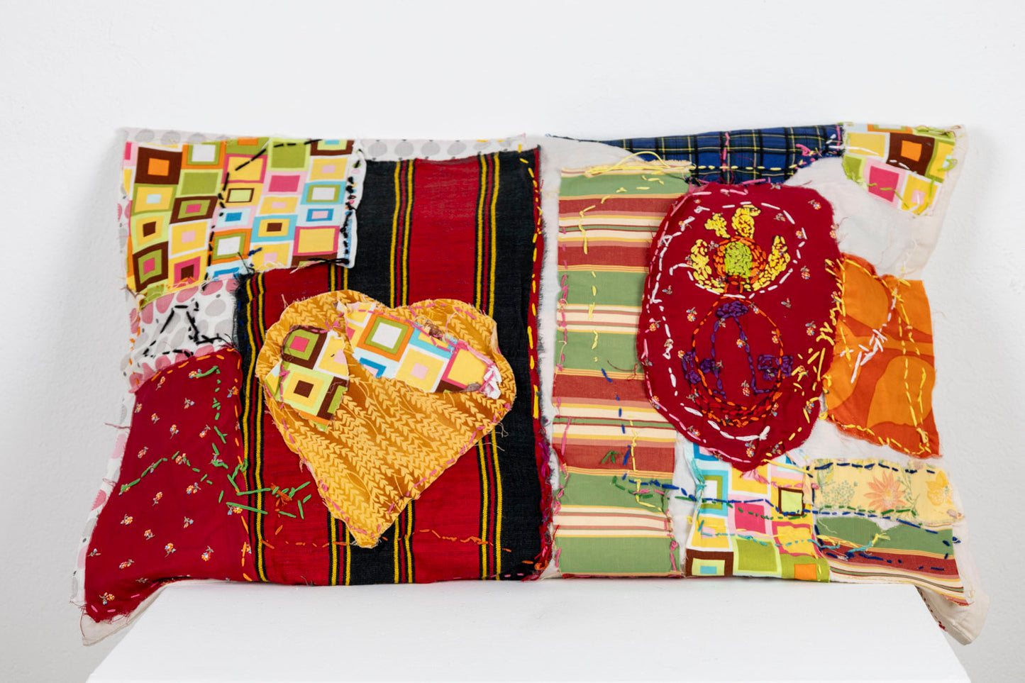 A bed pillow made of various patterned fabric swatches and stitched appliqué shapes. Some fabrics are striped, while others feature bright rows of squares or plaid patterns. One brown and yellow swatch is cut in the shape of a heart. There is a rainbow colored running stitch meandering across the surface.