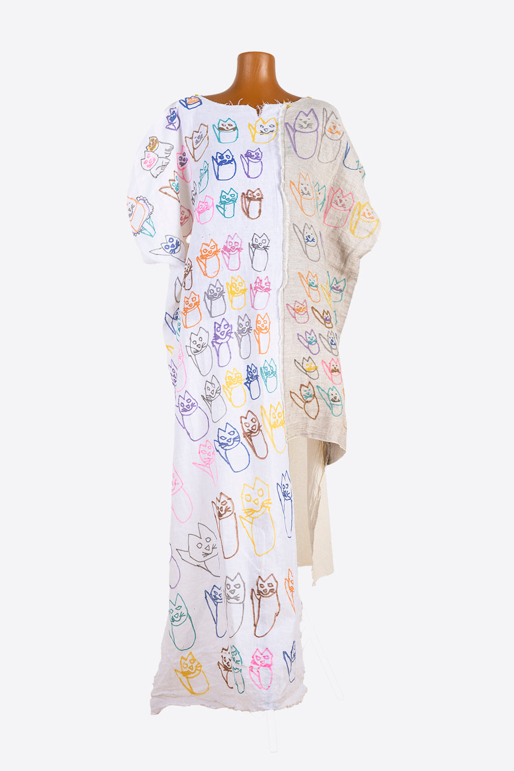 A long, hand-stitched kaftan, photographed on a wooden manikin. Two long panels of fabric are arranged side by side, and appear to be a gauzy white cotton and a thicker, tan linen. The white cotton extends to the ground and the linen side is cropped to knee-length. The artist has drawn rows of rainbow colored cats all over the garment.