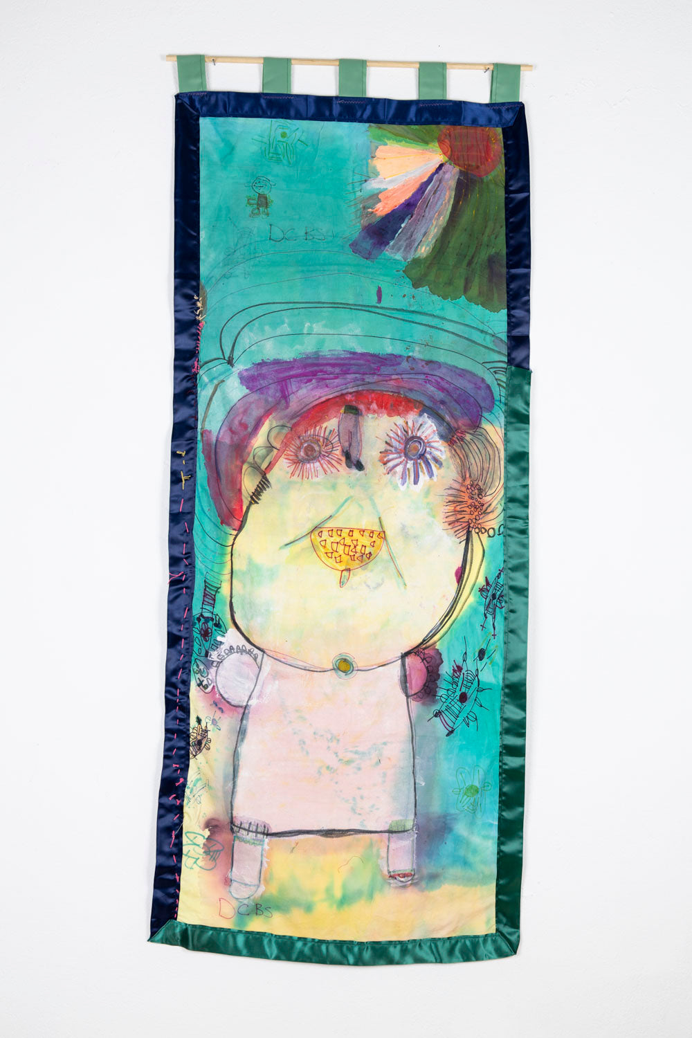 A wallhanging, mixed-media painted textile. The textile is a tall rectangle of tie-dyed fabric, mostly turquoise, with a shiny blue and green ribbon border. The artist has painted a large pink and yellow figure with a toothy grin, wide eyes framed by long lashes, and purple hair. Surrounding the figure are smaller designs, some of which look like butterflies or flowers. A red circle is tucked into the top right corner, with green and yellow rays radiating out around it.