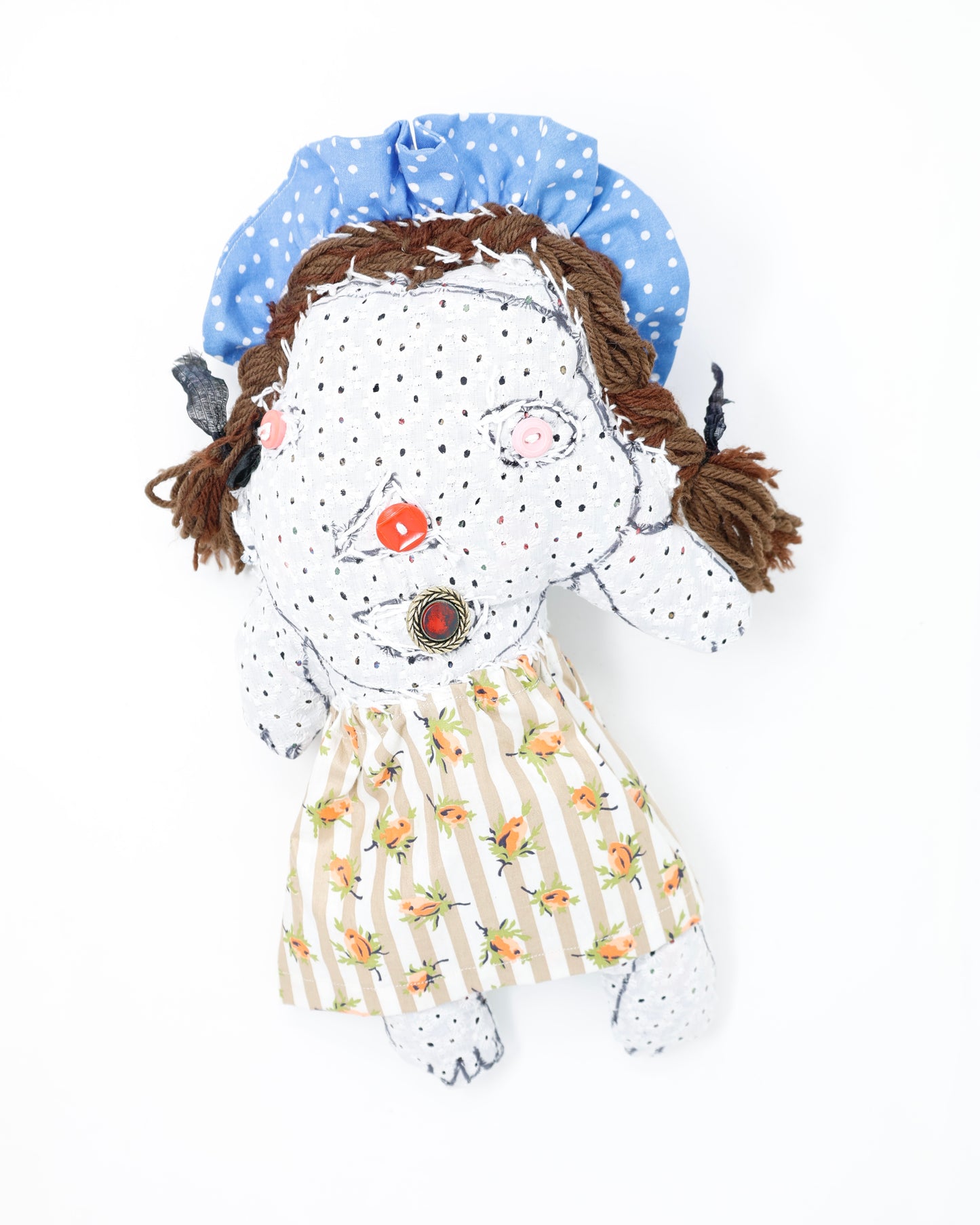 A fabric doll sculpture with a blue head piece with white polka dots. The doll is also wearing a beige and white striped garment with orange flowers on it. The hair is brown and braided, and the body of the doll is made with a light grey floral fabric.