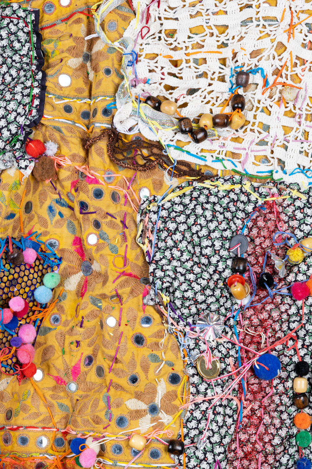This is detailed shot of a fiber art piece. The piece has a collage-like quality, with various fabrics and materials overlapping and layered. There is a black and white floral fabric alongside a white lace fabric layered on top of the yellow floral fabric. There are also pompoms and multi-colored threads.