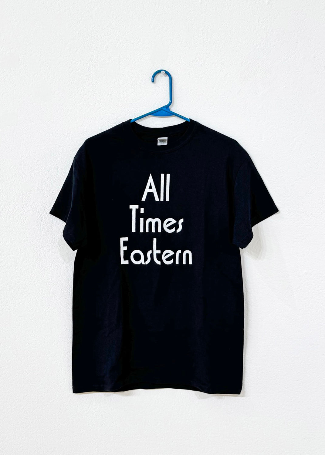 Black t-shirt with 'All Times Eastern' text on a white background, hanging from a blue hanger