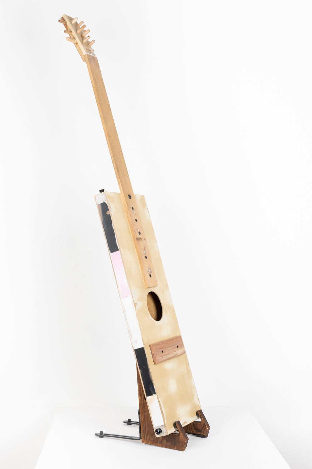 A handmade instrument photographed on a stand on a white pedestal. The instrument is a guitar made from reclaimed wood and metal. The body of the guitar is long and rectangular, and the tuning headstock is lined with little wooden pegs.