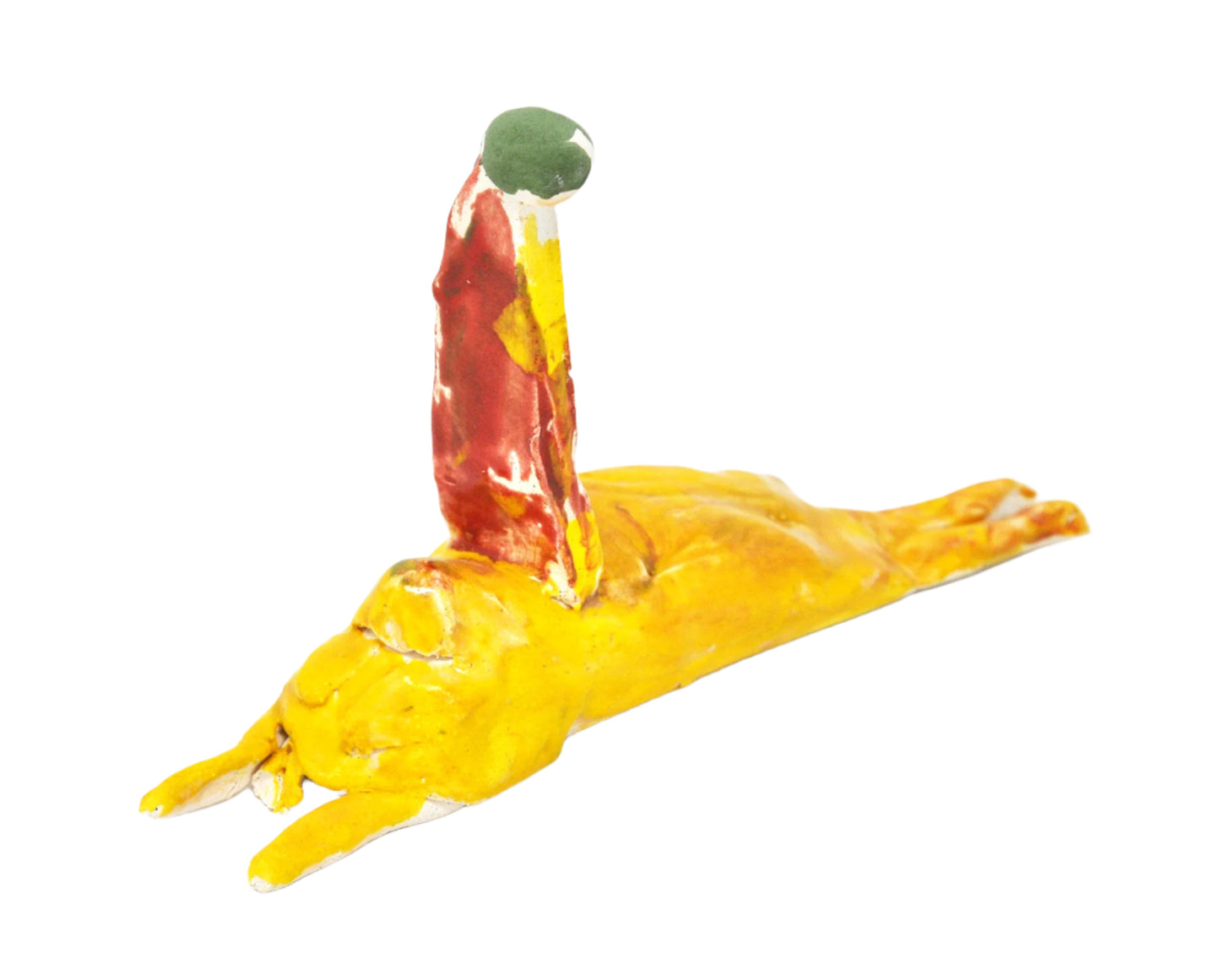A small ceramic sculpture of a dog wearing a red birthday hat. The dog is a long mound form, glazed bright glossy yellow and orange. It is lying down on its stomach with its thin legs sticking straight out from the front and back ends. A flat triangular hat with a round green pompom is positioned atop the dog's head.