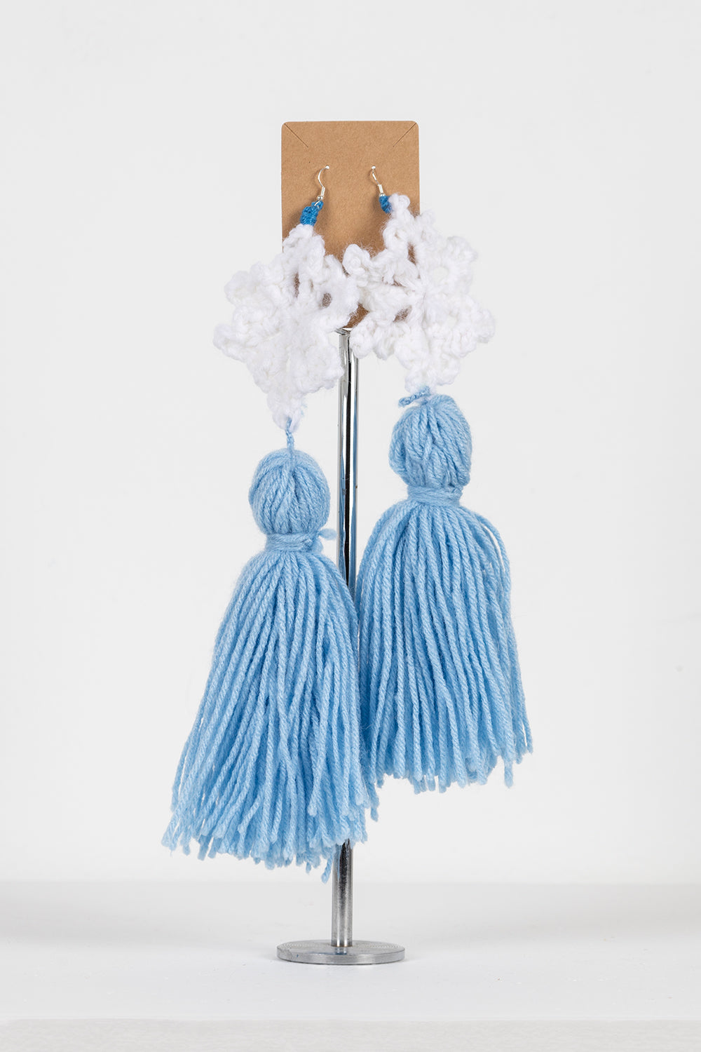 A pair of crocheted earrings hanging from on a tan card, photographed against a white background. The earrings are white yarn snowflakes with dramatic large blue tassels. They have thin silver hooks.