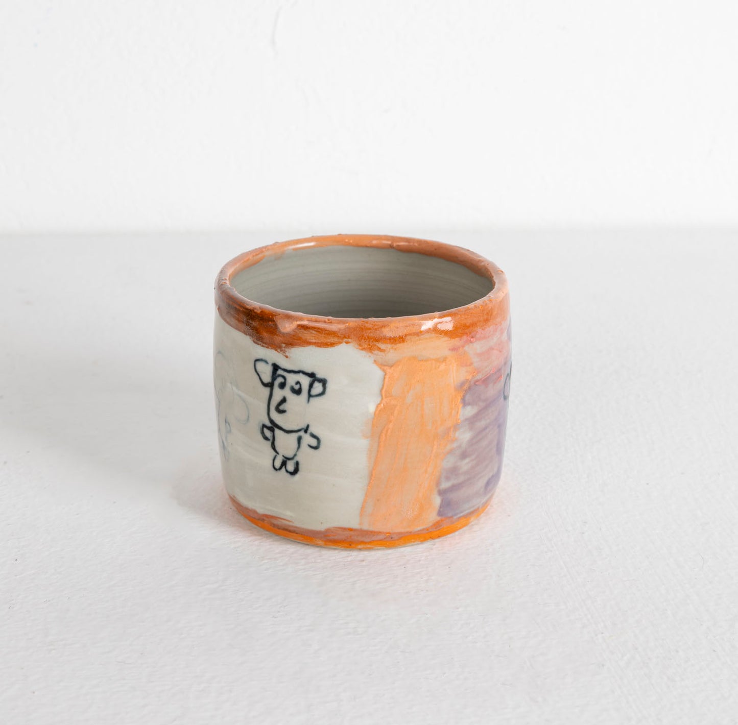 A small wheel-thrown ceramic cup, glazed with glossy purple, white, and orange. There are little figures painted around the outer surface.