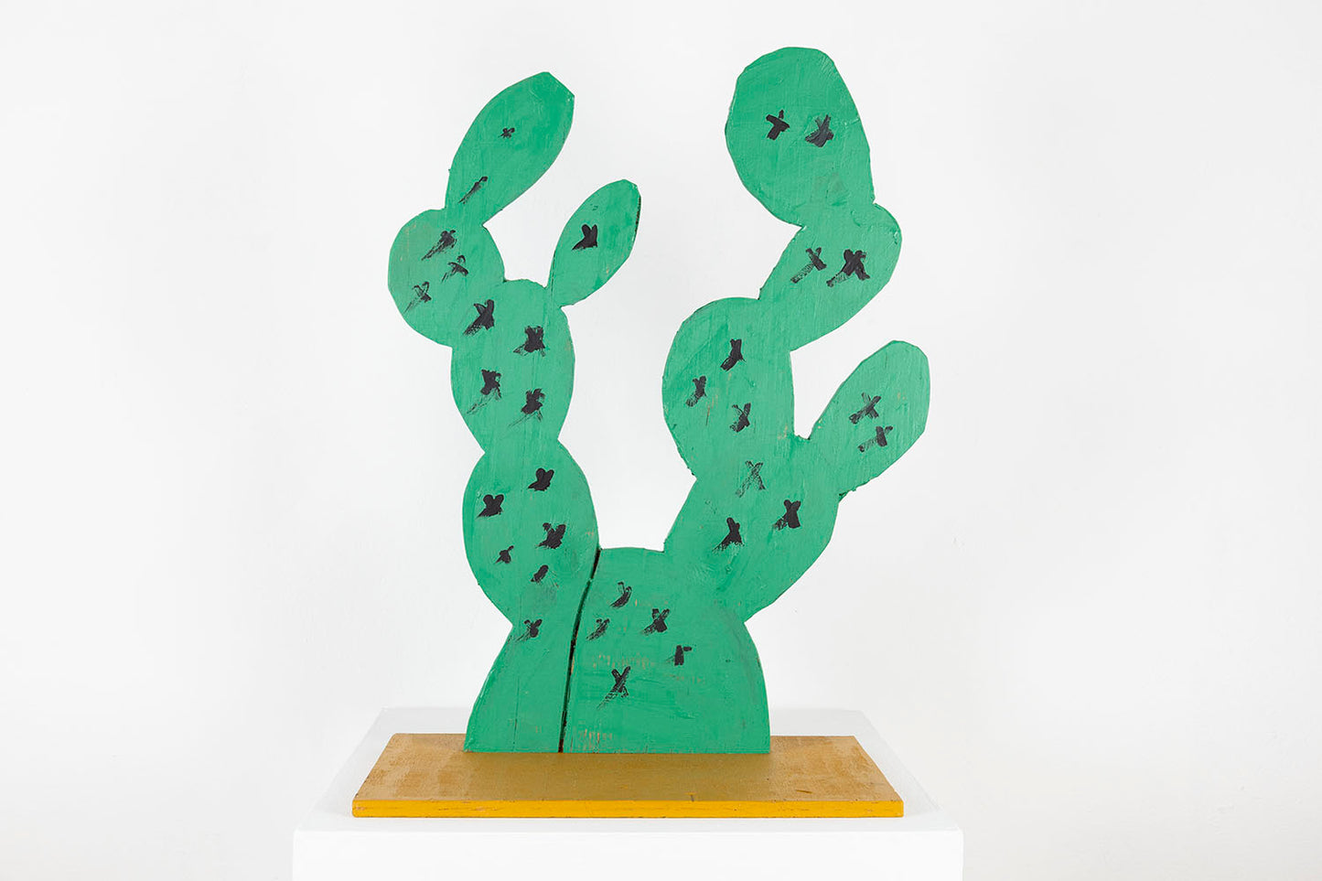 A wooden sculpture of a cactus, photographed against a white wall. The sculpture is made from flat boards of plywood which have been cut to mimic the silhouette of a lumpy cactus with two spiny branches extending straight up. The wood is painting bright green with little black X-forms representing thorns speckled all over. The cactus stands upright on a square yellow wooden base.