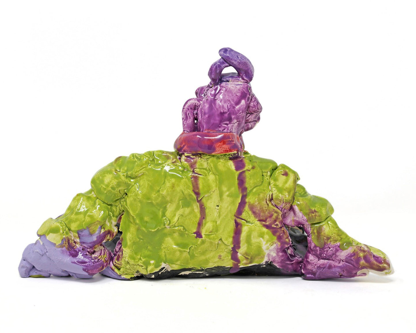 This ceramic sculpture depicts a figure that is sitting down. The figure has purple skin, and is wearing a green shirt with yellow buttons, a red scarf, black pants and brown shoes.