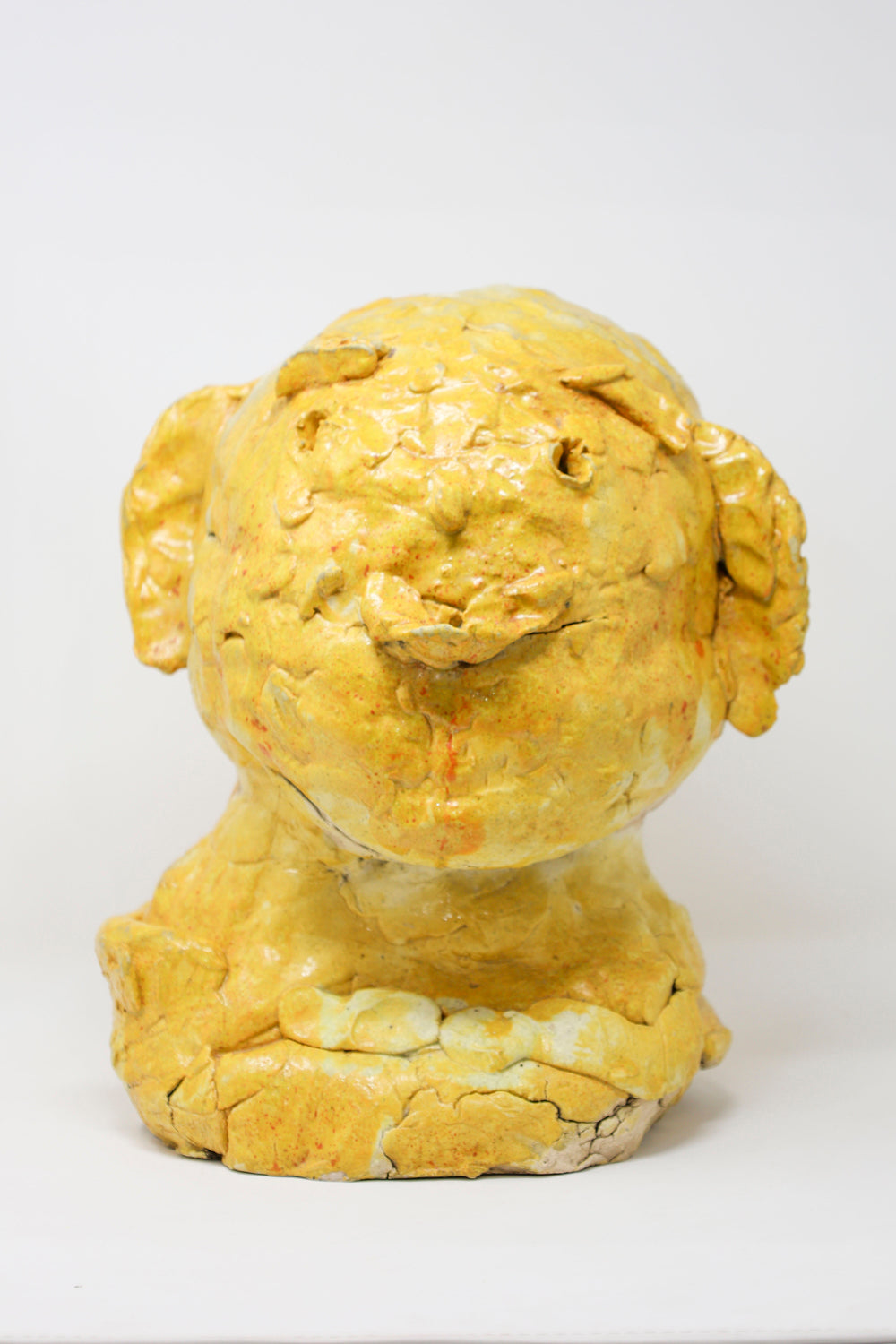 A highly textured yellow ceramic sculpture of a human head, against a white background. The person is facing forward, and has a stoic expression.