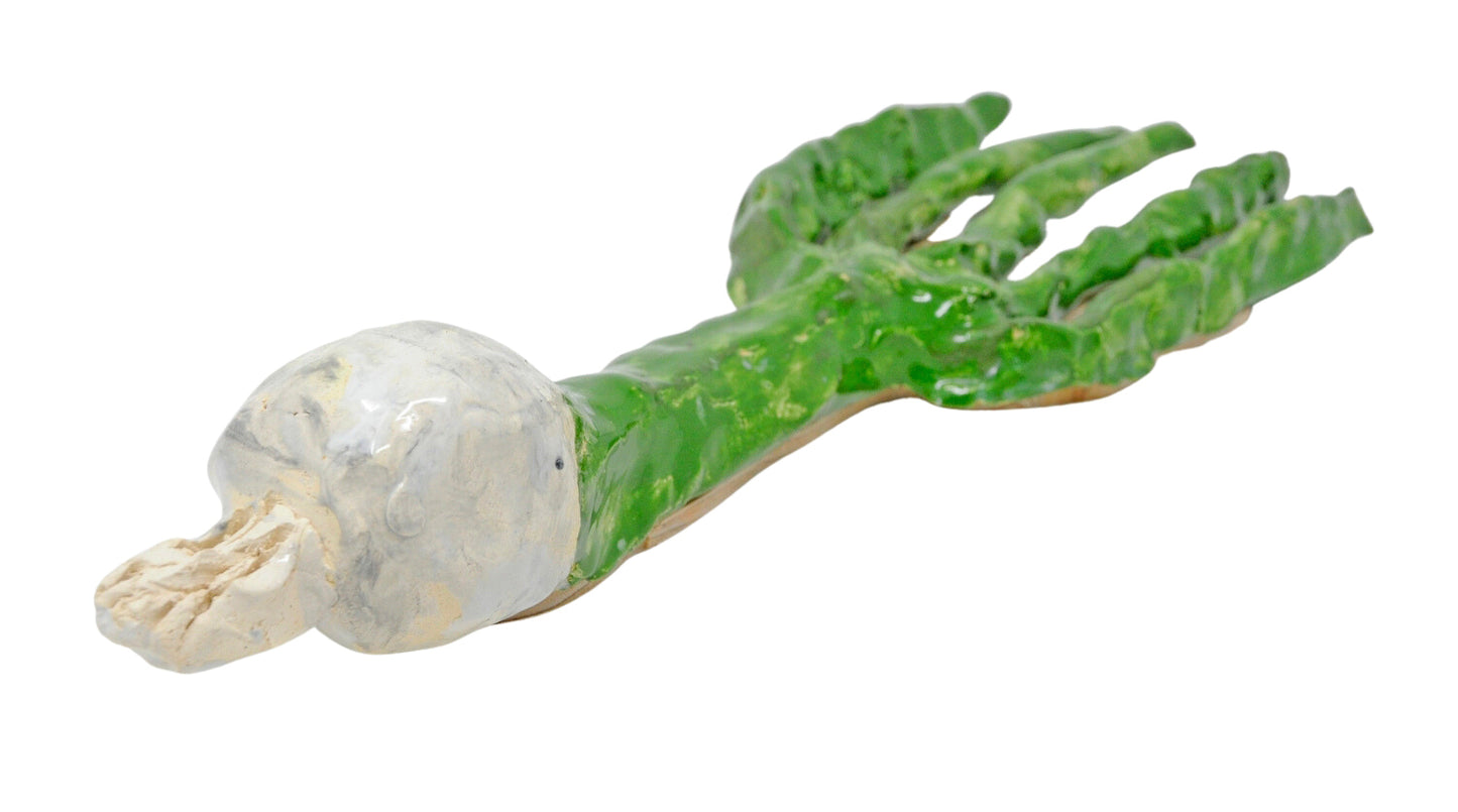 The sculpture photographed from a different angle, with the white root bulb in sharp focus in the foreground. The full description from the previous image describes this item as a ceramic sculpture in the shape of a green onion vegetable, glazed green and white. The object is a long green tube that branches out into 5 green stalks on one end. The other end of the sculpture is a small white colored bulb, representing the root bulb of the onion.