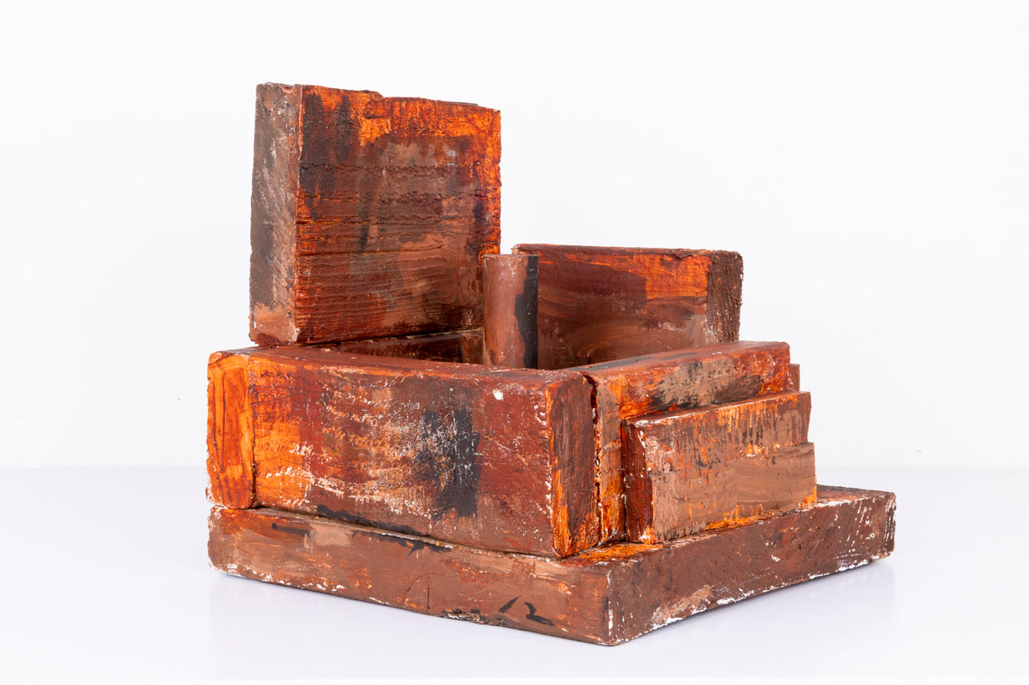 An abstract sculpture made of stacked rectangular wooden blocks painted in layered shades of reddish brown, copper, and black. The geometric forms are arranged in a compact architectural composition on a square base, with visible brushstrokes and rough textures emphasizing the handmade construction. The sculpture is displayed against a plain light gray background.