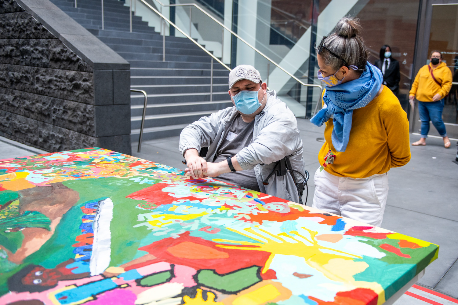 A photograph of two people in a museum courtyard working on a large painting. The figure in the center is man with light skin who is seated in a walker. He wears a grey baseball cap, a blue face mask, and a grey jacket. A white woman with grey hair in a bun stands to him. She is wearing a blue scarf, yellow sweater, and white pants. A large painting with green, yellow, and red shapes is laid out flat on a table in front of them.