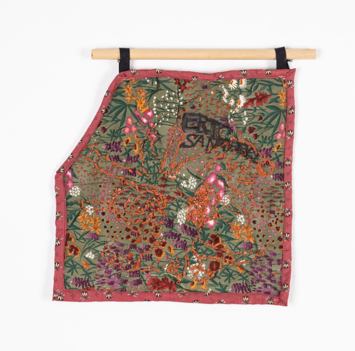 A shaped wallhanging textile work hanging from a wooden dowel and photographed against a white backdrop. The main swatch of fabric is printed with an image of a garden: pale green ground with yellow, white, red, and purple flowers. The fabric is cut like a square with one corner folded down. Over this design, the artist has embroidered a swirling shape using orange thread and written words including "Eric" with black fabric marker. The piece has a dark pink floral fabric border.