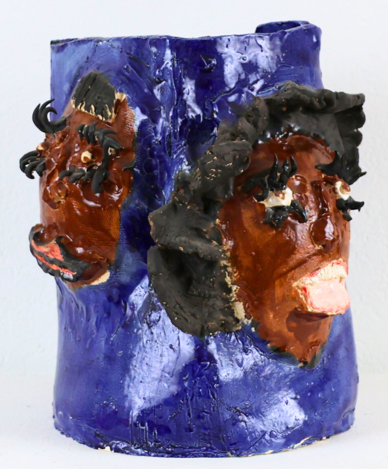 A tall ceramic vase featuring sculpted portraits protruding from the sides. The portraits are glazed brown with long curled eyelashes and scalloped black hair. The portraits represent Aretha Franklin. Except for the faces, the entire vase is painted in a shiny royal blue glaze.