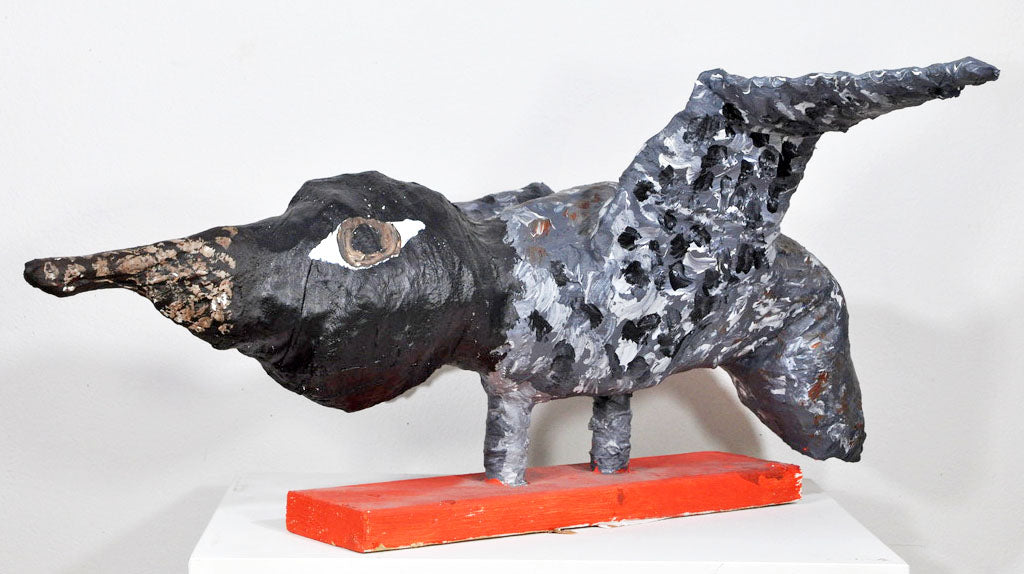 A papier mâché sculpture of a speckled black, white, and grey bird perched on a red wooden board. The bird has a large brown and white eye and a pointy black beak dotted with copper paint. Its wings are spread wide as if it's about to take flight.