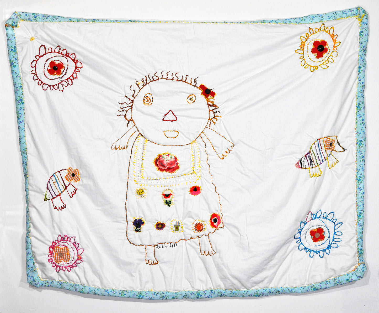a wall hanging quilt with a light blue border and various figures stitched on the surface. The central stitched image is a large figure. There are also four flowers in each corner and two birds on each side of the figure.