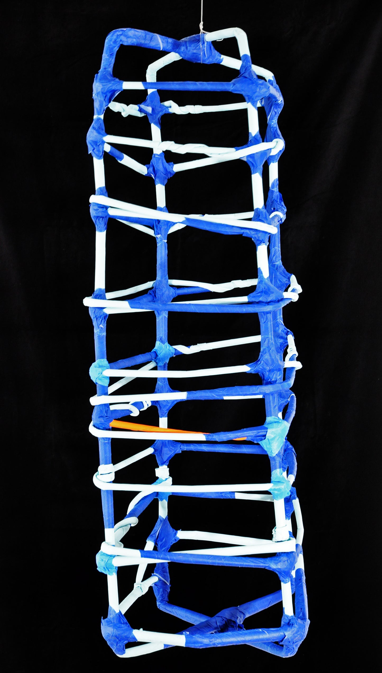 a sculpture made of tightly rolled tubes of paper and blue painters tape, photographed against a black backdrop. The form is a dimensional grid or cage-like structure. The white paper tubes are bound by blue tape wherever they intersect.