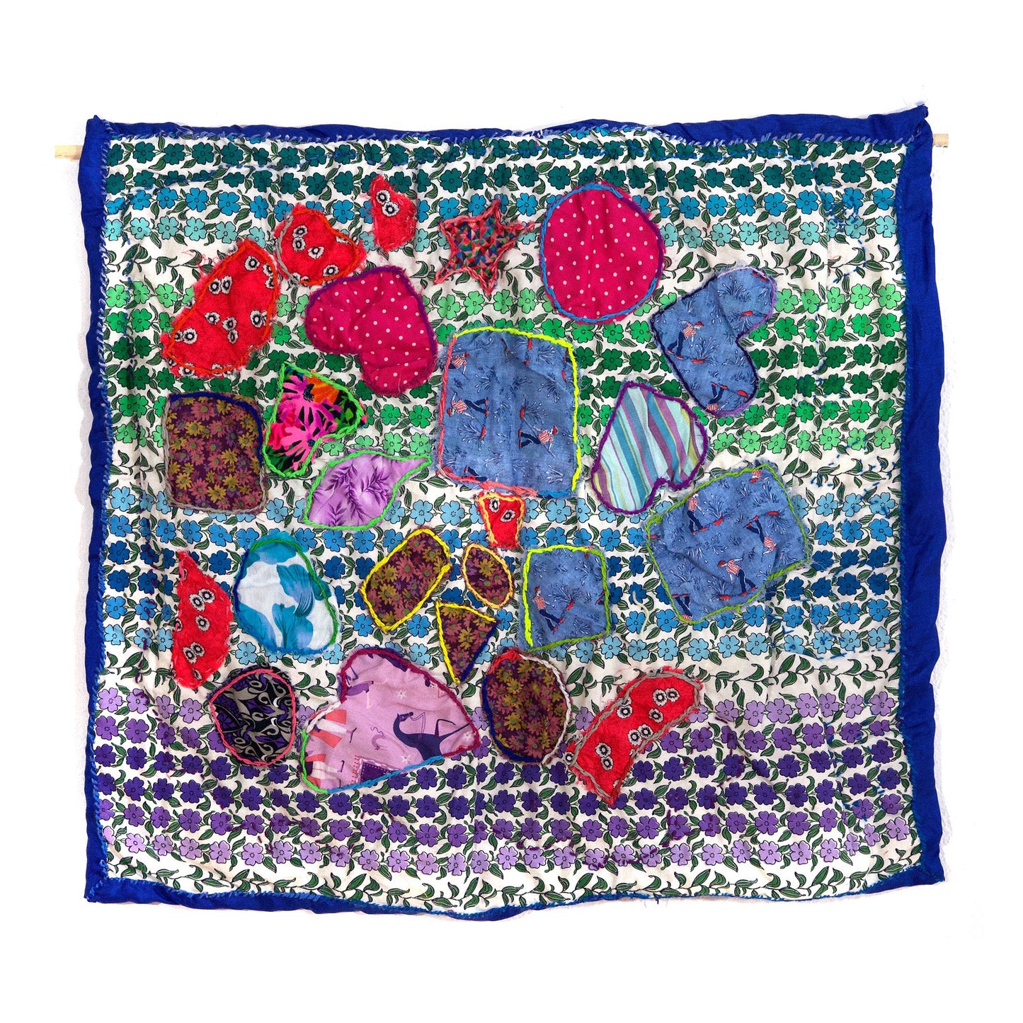 An embroidered fiber wall hanging. The main square of fabric is white with rows of blue, green, and purple flowers printed over its surface. There is a dark blue border stitched around the perimeter. The artist has cut hearts, circles, and squares out of various pieces of red, blue, and pink patterned fabric and stitched them on top of the larger square. A rainbow thread running stitch is visible around each shape.