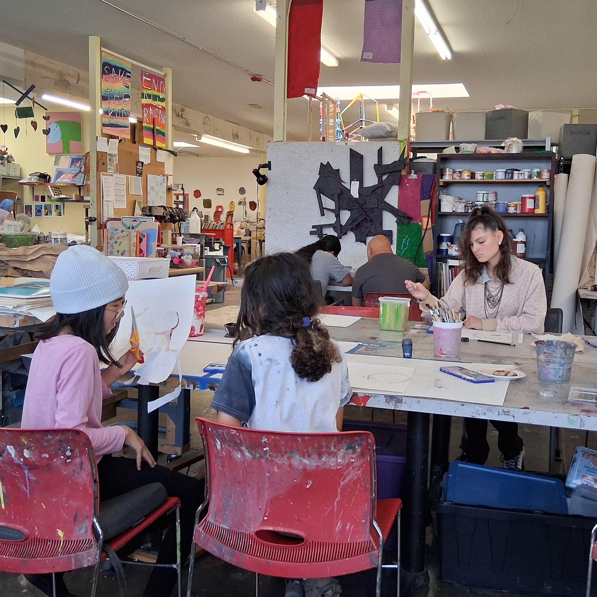 People work at tables in a vibrant art studio surrounded by colorful paintings, supplies, and paint-splattered red chairs.