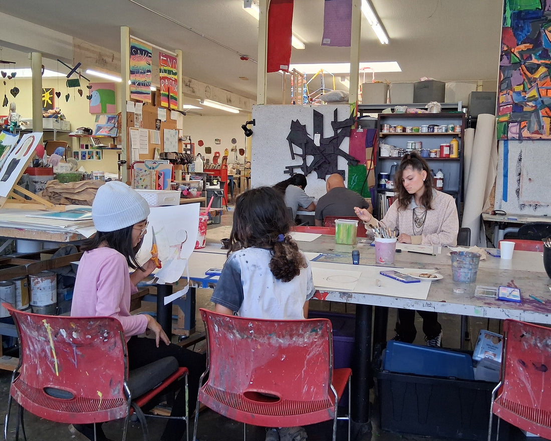 People work at tables in a vibrant art studio surrounded by colorful paintings, supplies, and paint-splattered red chairs.
