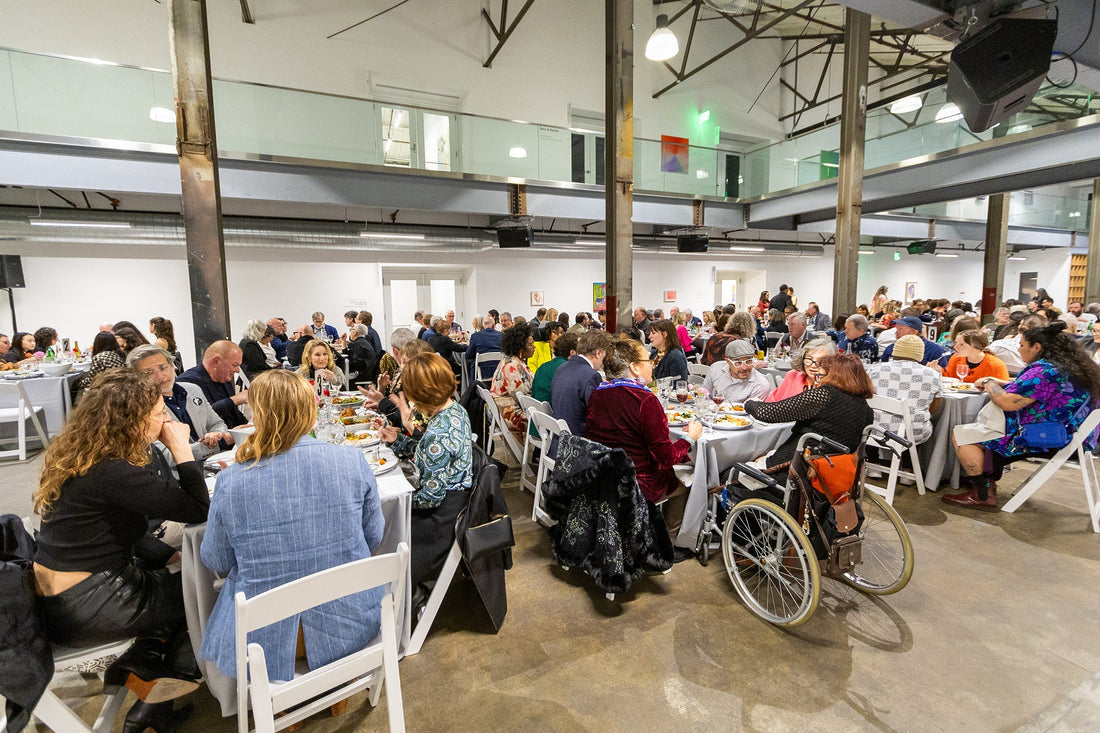 Guests gather at round tables covered in white tablecloths throughout NIAD's industrial art studio space during the 2025 Artful Gala fundraiser. Attendees in colorful attire enjoy their meals while surrounded by artwork displayed on the gallery walls.