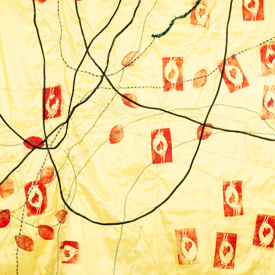 Detail of a large square of yellow satin mixed media artwork. Synthetic hair braids loop and intersect across the surface, stitched down with green thread. Lino-block stamps with red ink shaped like petals or eyes. 