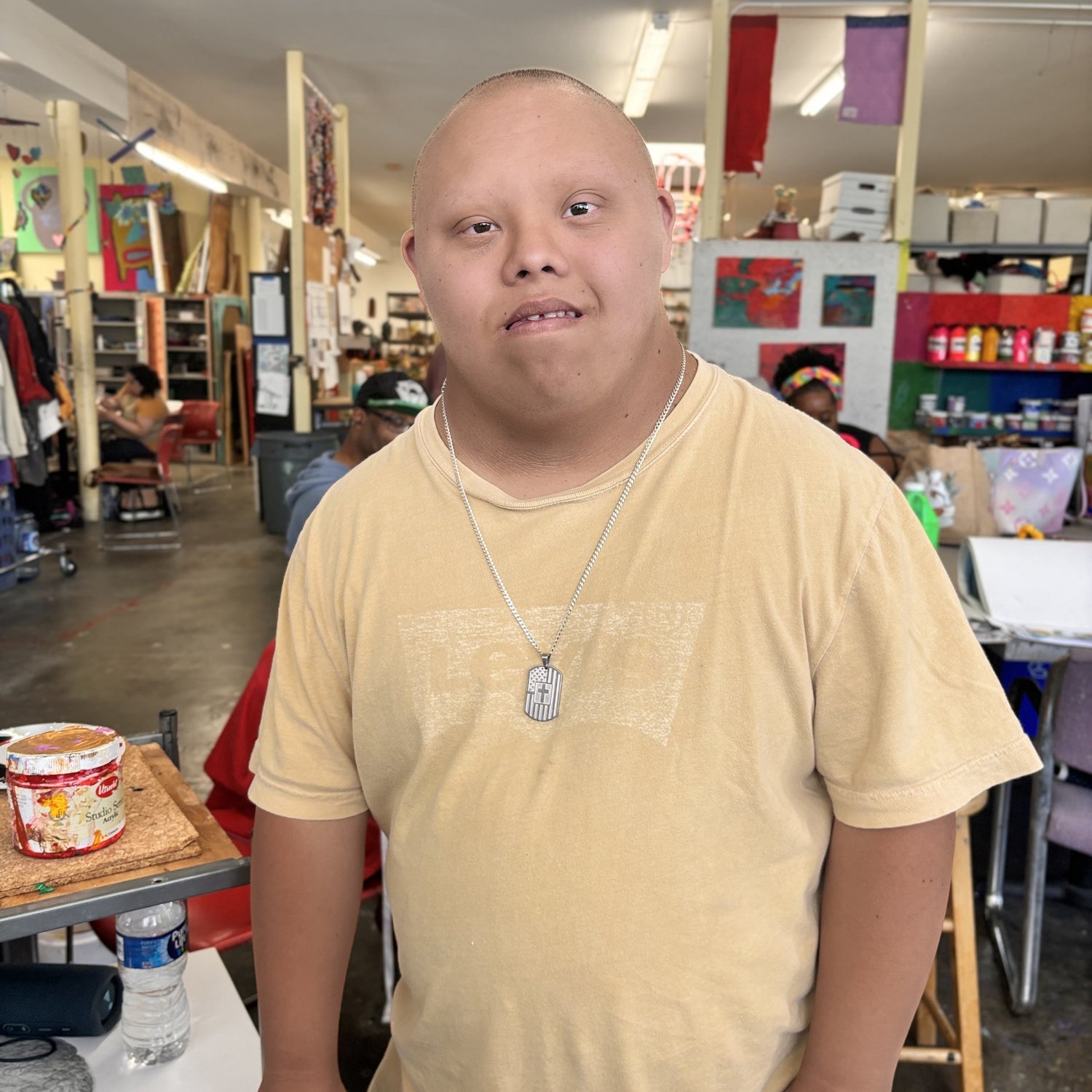 in an art studio setting, a person with close-cropped hair and light brown skin wearing a dog tag necklace and tan shirt, looking straight at the camera, posing next to their art work area.