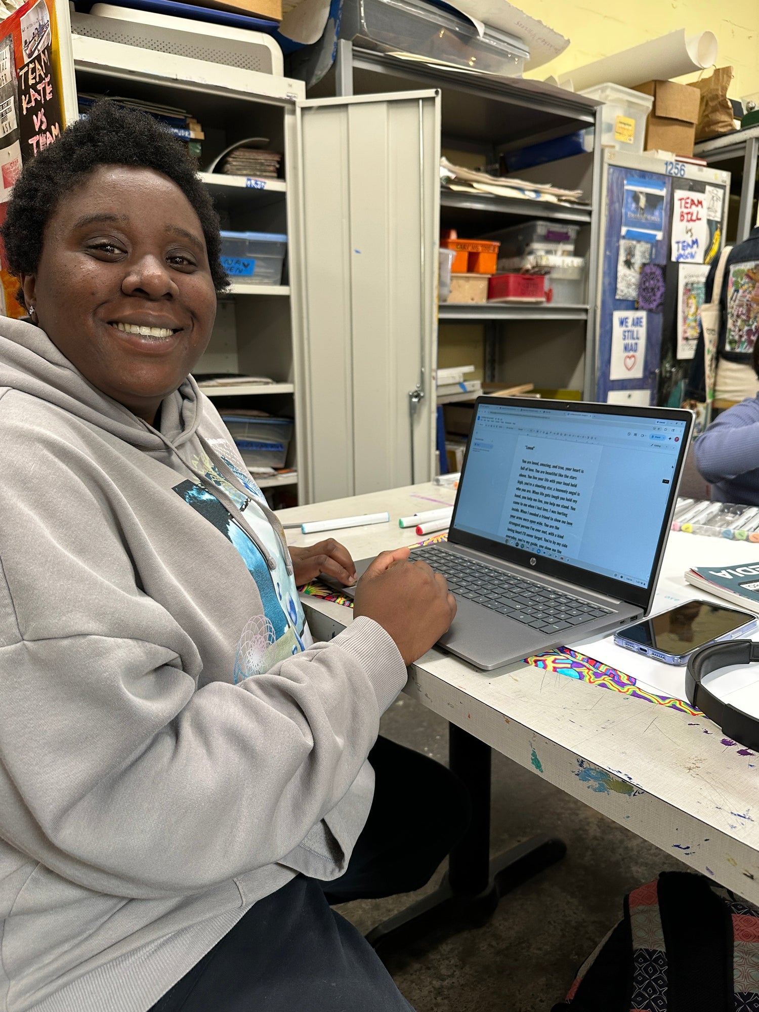 a black woman with short hair wearing a grey sweatshirt. She is smiling and sitting at a desk writing on her laptop. 