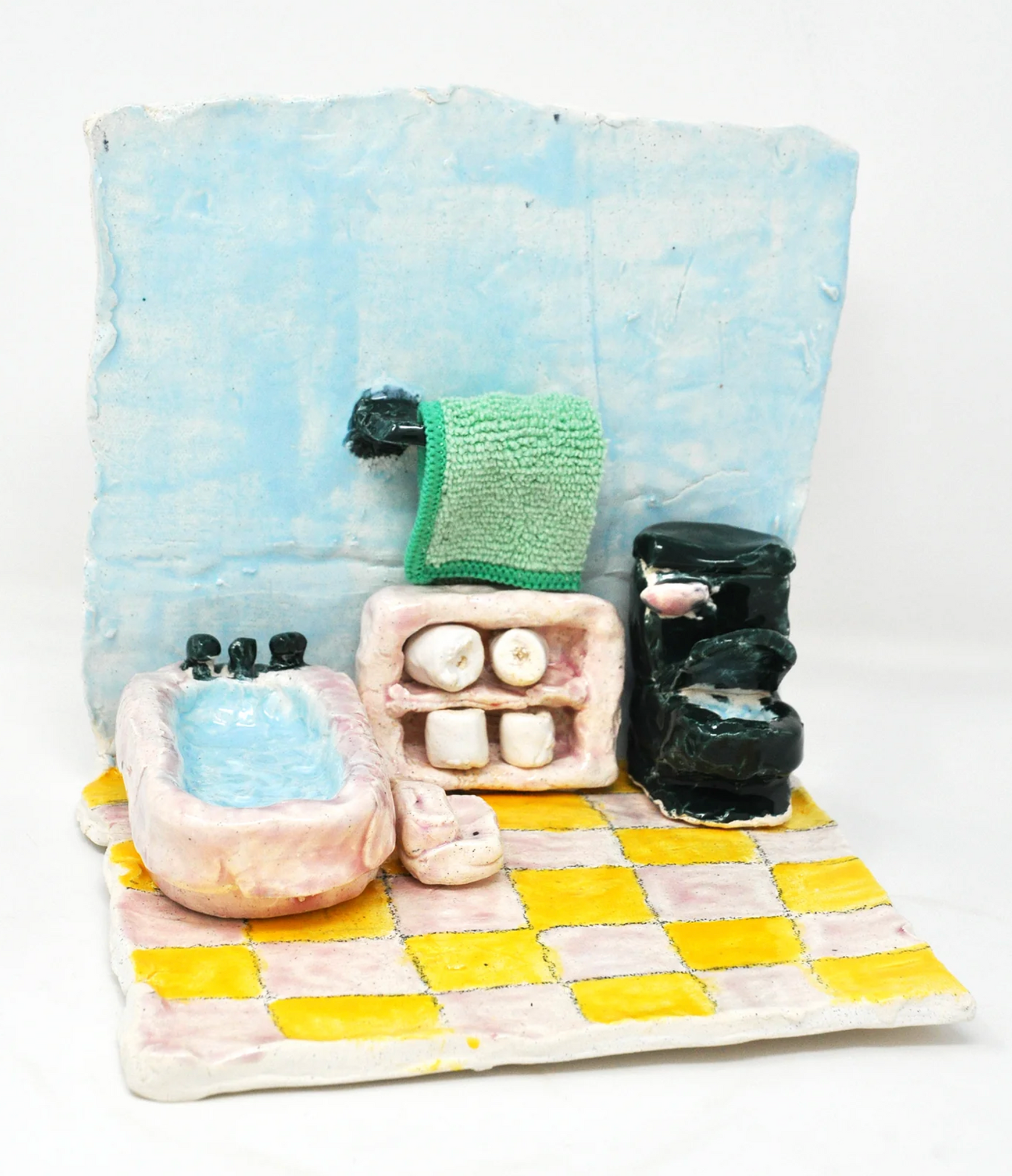 A miniature diorama of a bathroom made of glazed ceramic. The floor of the scene is tiled in a yellow and pink grid. The items in the room include a pink tub full of blue water, pink shelves, a fuzzy green towel, and a black toilet. 