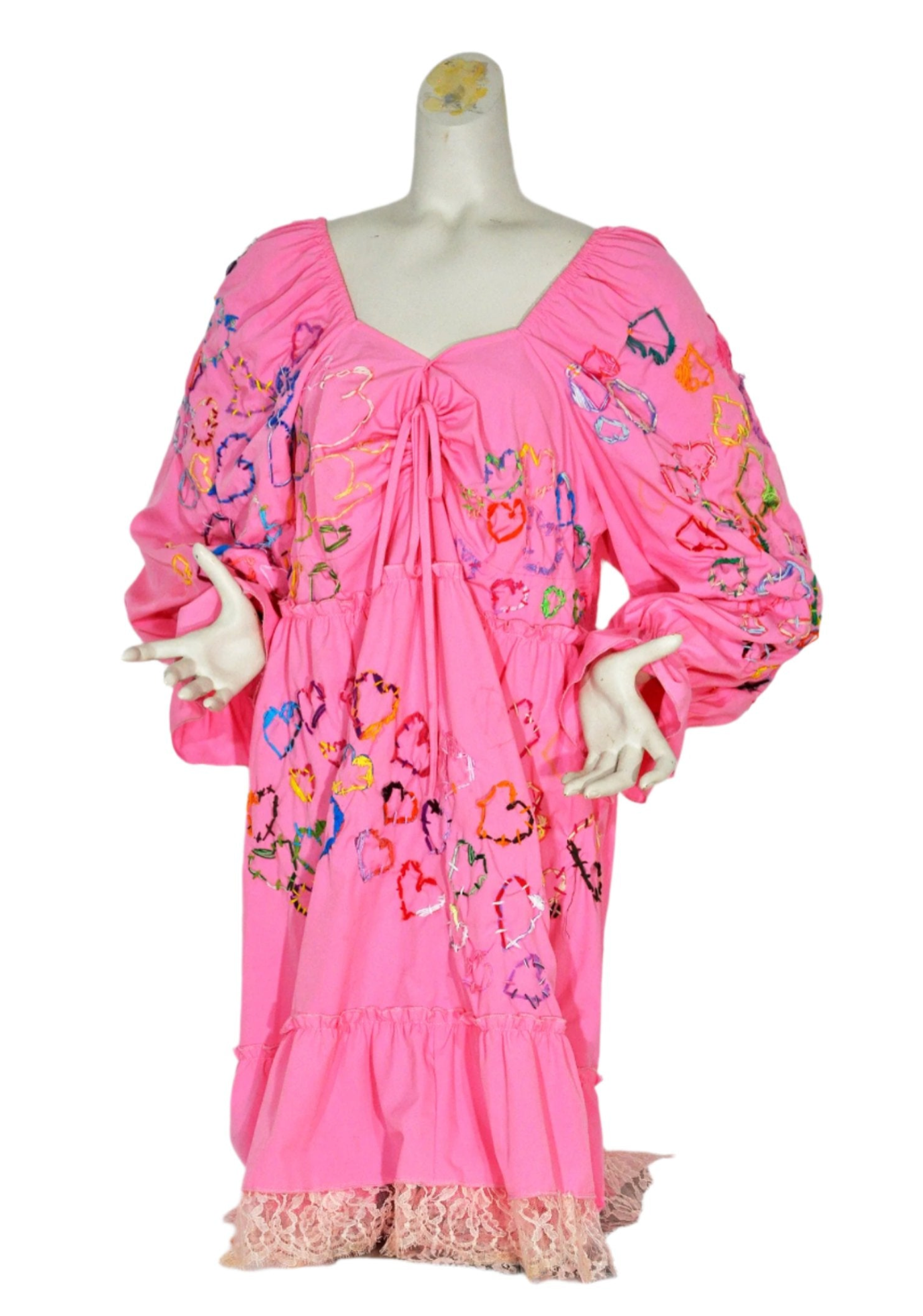 a pink long sleeve dress modeled on a grey manikin torso. The artist embroidered hearts in every color of the rainbow across the front and sleeves of the dress. There is pale pink lace trim stitched along the bottom hem.