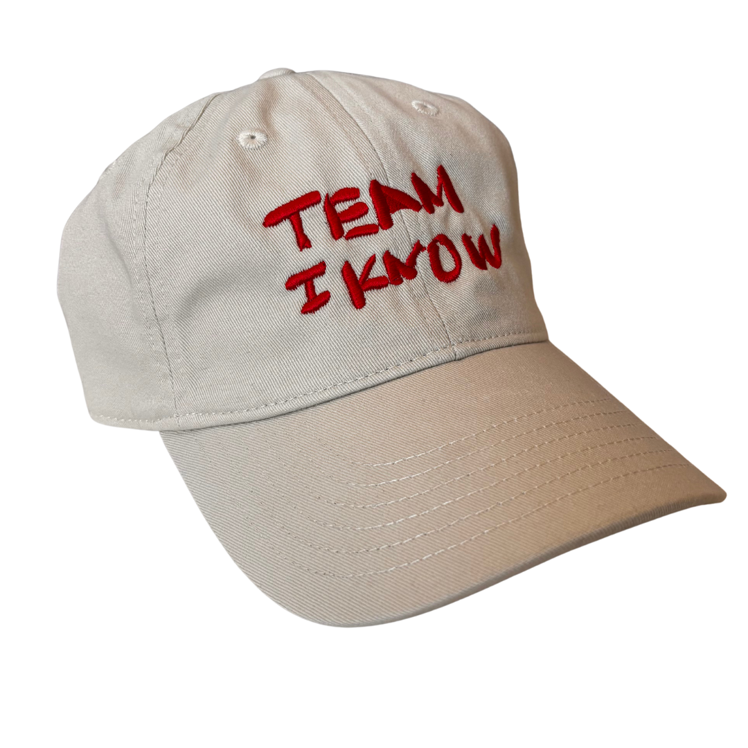 Light khaki-colored baseball hat with the words "TEAM I KNOW" embroidered in red on the front in a handwritten font. The hat floats in a white void.