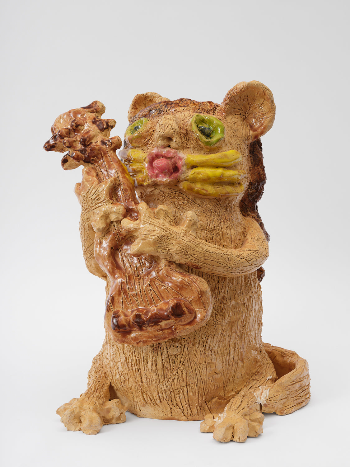 A tan ceramic cat with many textured lines delineating fur is seated on its hind legs grasping an acoustic guitar with its hands. The cat has big green circular eyes, fat yellow whiskers, and a pink mouth with the cat's tongue sticking out. 