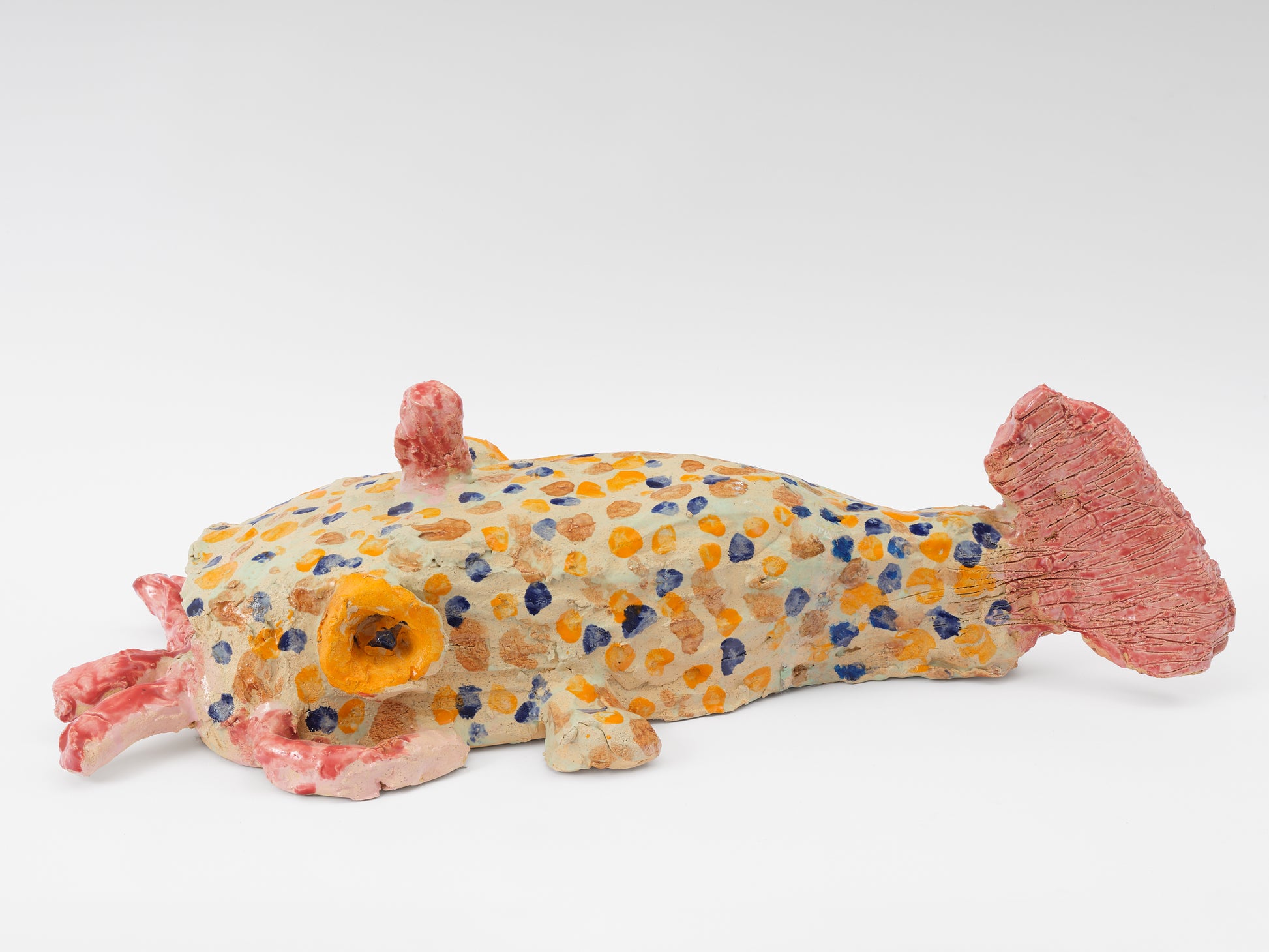 A sculpture of a ceramic catfish, pictured from the side. The large fish has birght orange and blue spots all over its pale yellow body. It has a bright pink tail and thick whiskers that are the same hue. The fish has a large orange circular eye.