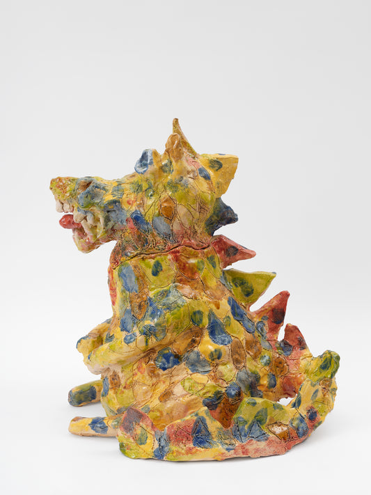 A profile view of a ceramic yellow dragon sculpture that is dappled with blue, ochre, and soft red glaze all over. The rotund dragon has spikes going down its back, and its tongue sticks out of its mouth