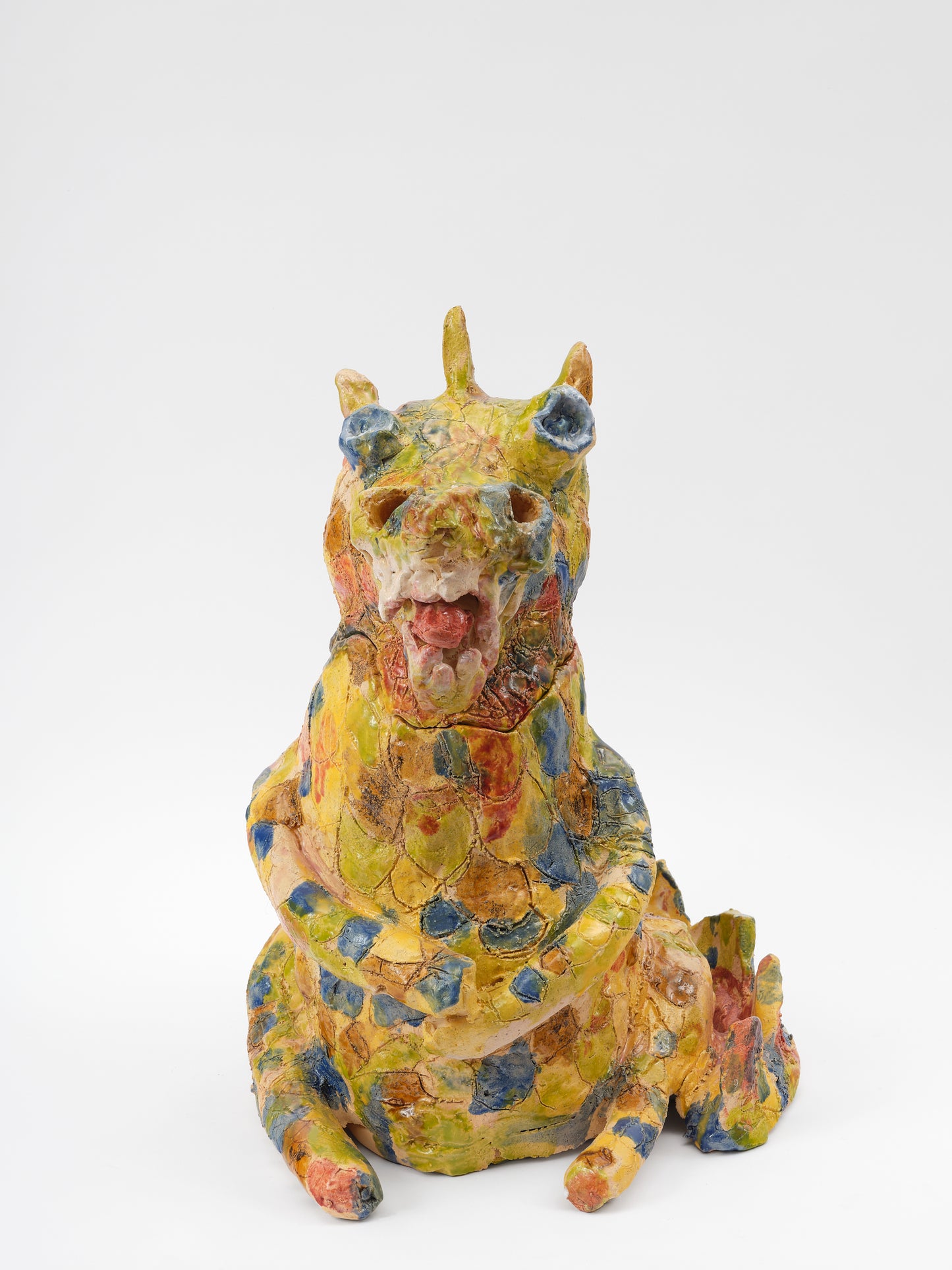  A ceramic yellow dragon sculpture that is dappled with blue, ochre, and soft red glaze all over. The rotund dragon is seated and has its arms crossed. The dragon's mouth is open and is showing large teeth and sticking out its tongue. 