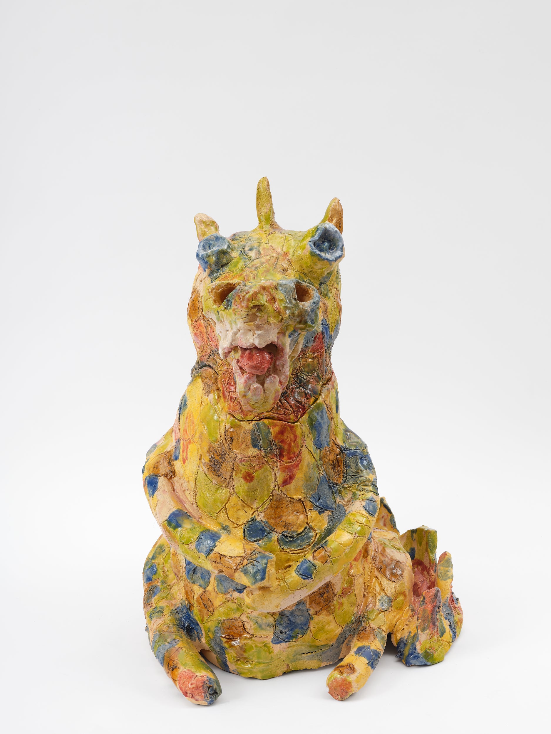  A ceramic yellow dragon sculpture that is dappled with blue, ochre, and soft red glaze all over. The rotund dragon is seated and has its arms crossed. The dragon's mouth is open and is showing large teeth and sticking out its tongue. 