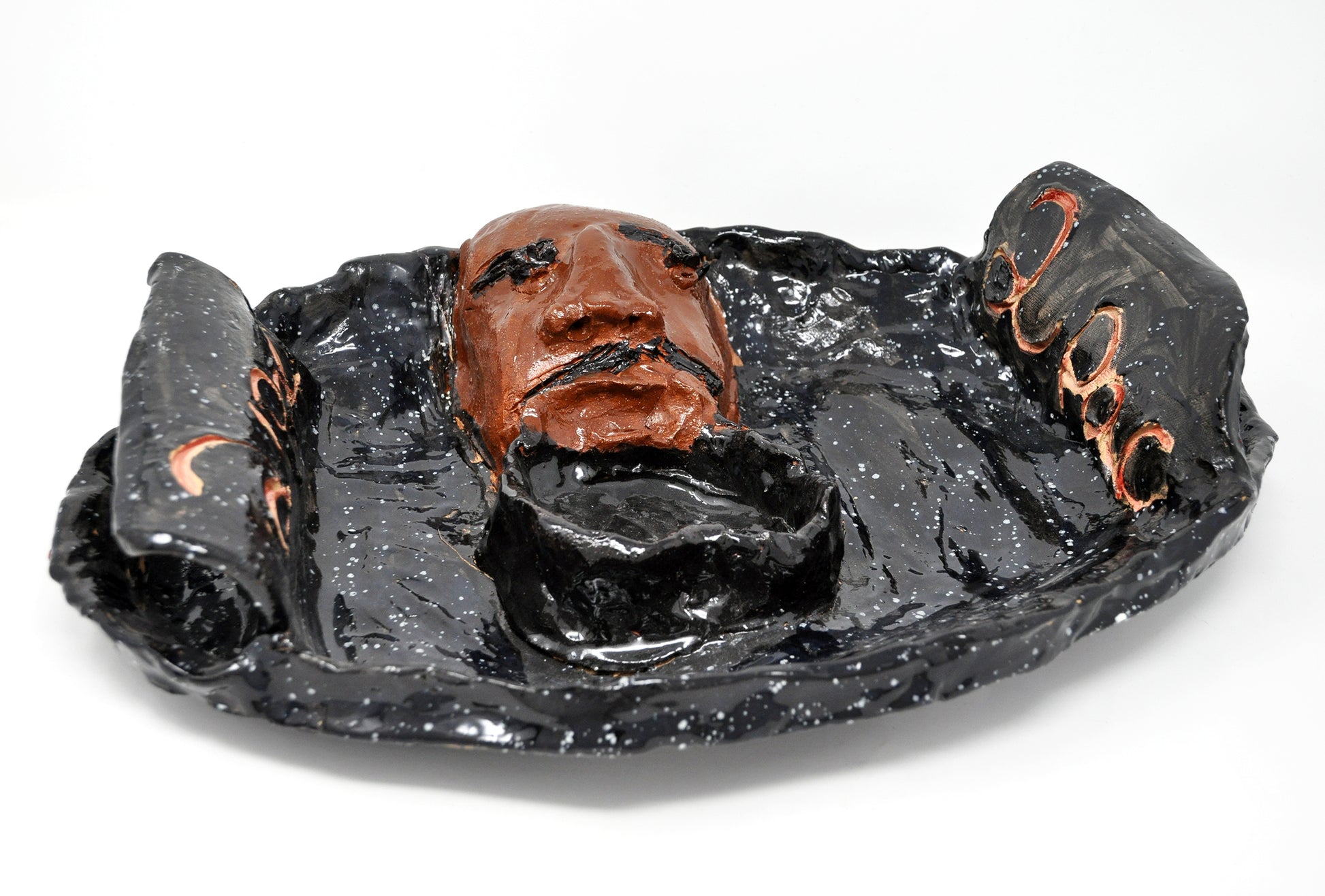 A glazed ceramic platter with a sculpted portrait in the center. The portrait represents the musician 2Pac. The head is painted brown and has black eyebrows and a black mustache. There is a shallow black bowl form connected to the lower portion of the sculpted head. On either side of the platter is a curved slab handle, both etched with the name "2Pac". The platter is painted glossy black with white speckles, like a night sky full of stars.  