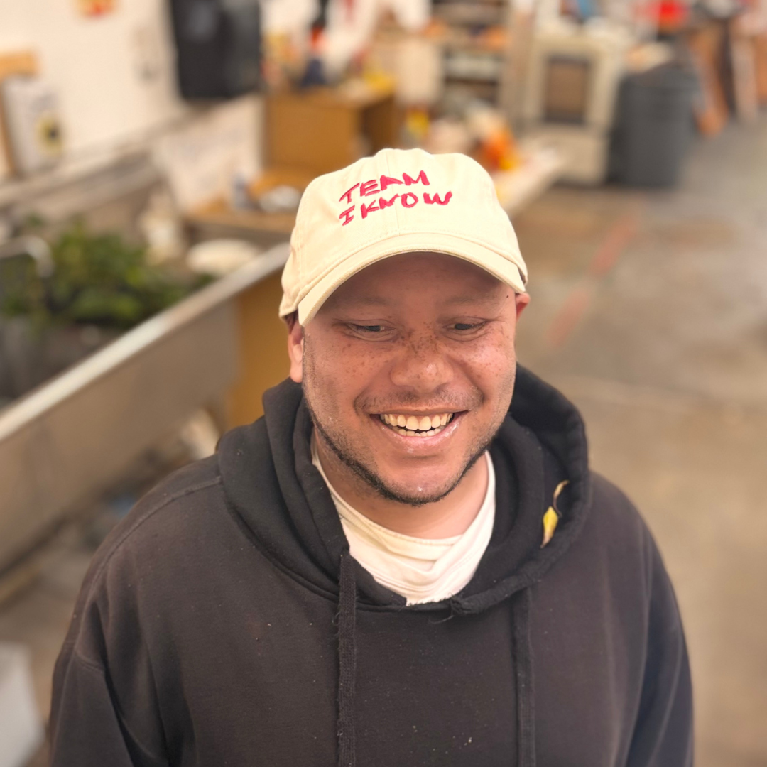 NIAD artist Jason Powell Smith - a man with light brown skin in a black hoodie, smiling widely - modeling the hat that features his "Team I Know" artwork. Jason stands in an art studio setting, blurred in the background.