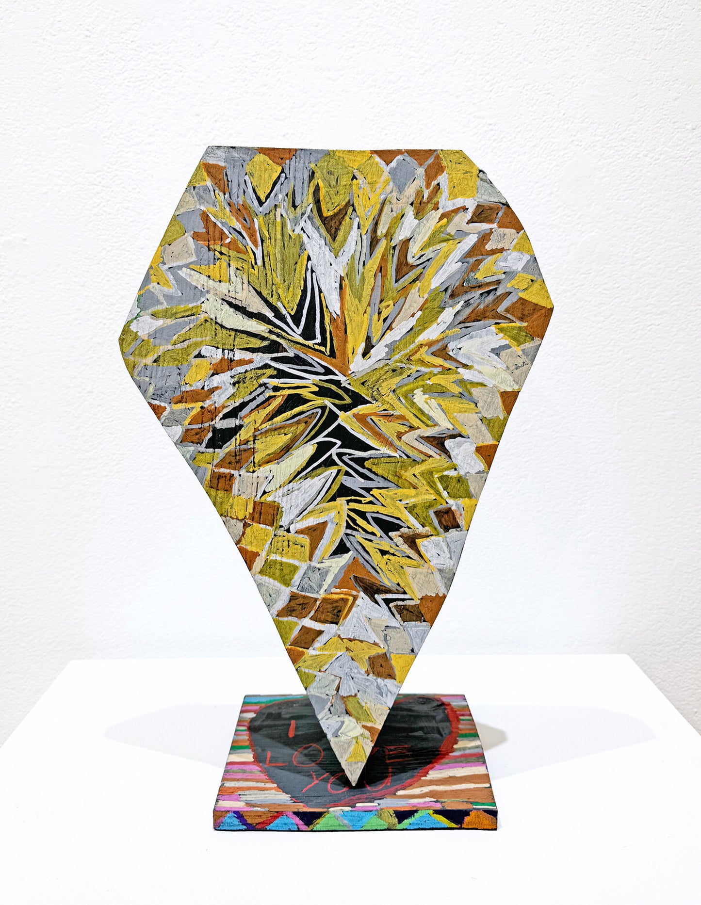 A diamond shaped wooden sculpture rests with the narrow tip on a small square wooden base. The base has bright colors of blue, green, red, and pink drawn in jagged patterns and lines along the edge, and there is a black form outlined in red on the top of the base that says "I Love You." The diamond shaped form has abstracted, spiky patterns that are in colors such as yellow, white, grey, and a rusty orange.  