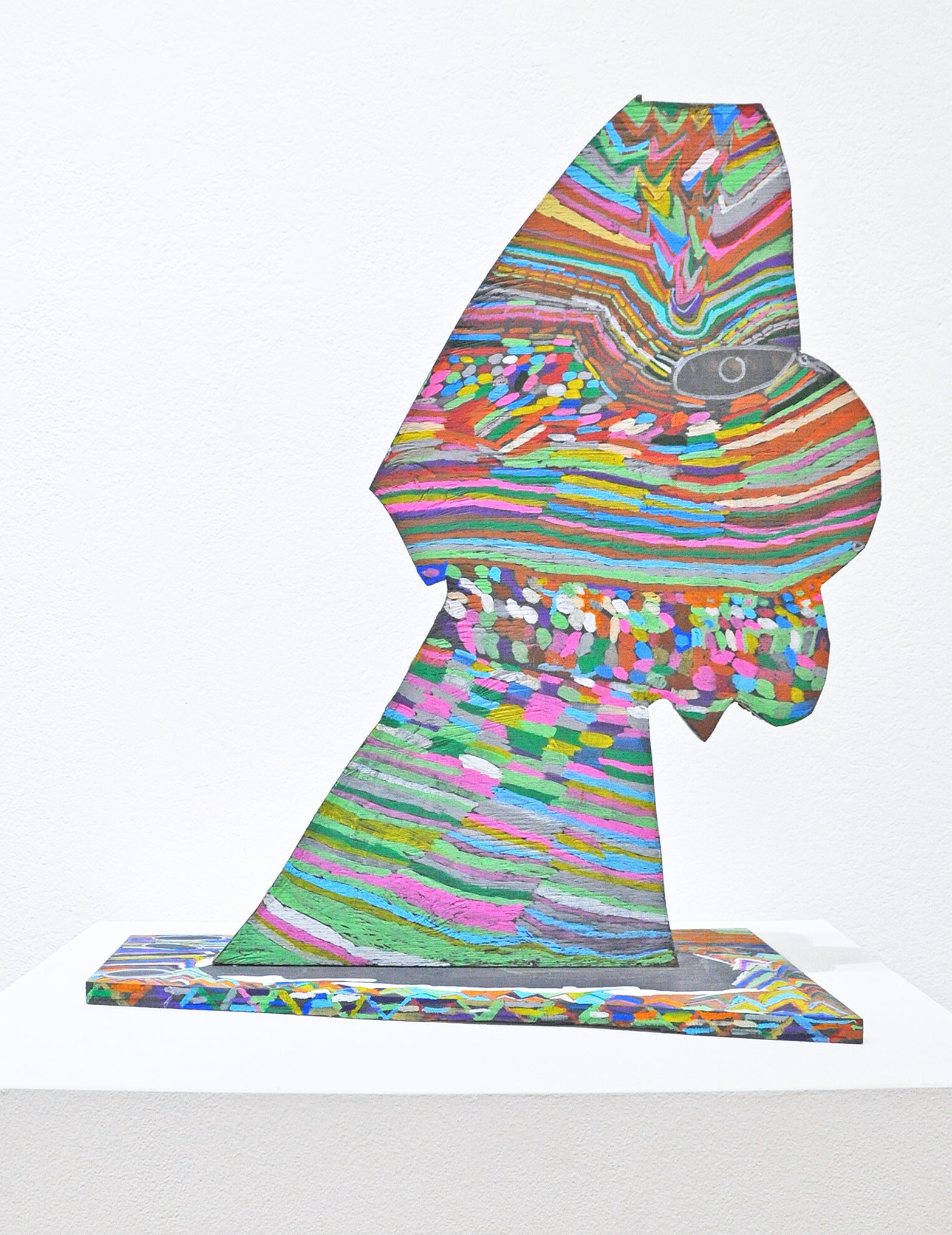 A wooden sculpture - a shaped plank, tilting to the right, affixed to a base plank. Both are embellished with bands of bright color, striated like the patterns left in rocks over millenia, with an almond shaped eye indicating that the shape might be a human figure.