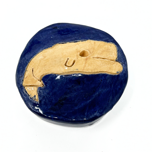 A sculpture photographed on a white surface. The object is a thick ceramic disk, glazed navy blue, with a drawing of a sperm whale etched onto the top surface. The whale is glazed a tan color and has a deep round hole for an eye.