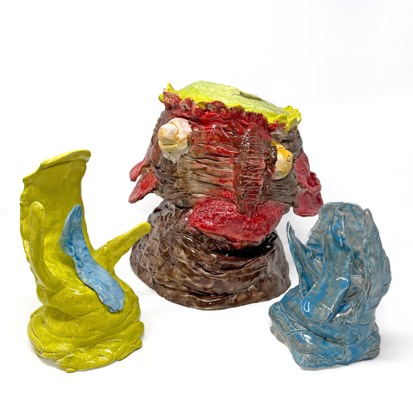 A 3-part ceramic sculpture of a portrait head and two hands. The sculpture represents the Swamp Thing monster from comic book and movie fame, a hybrid human fish creature. The sculpture is made of stacked clay coils, glazed brown, red, and yellow. The face has two bulbous white protruding eyes, big red lips, and 4 red flaps representing gils hanging on the side of the head. The hands look like gloves with three large webbed fingers. One hand is glazed bright yellow and the other is grey and blue. 